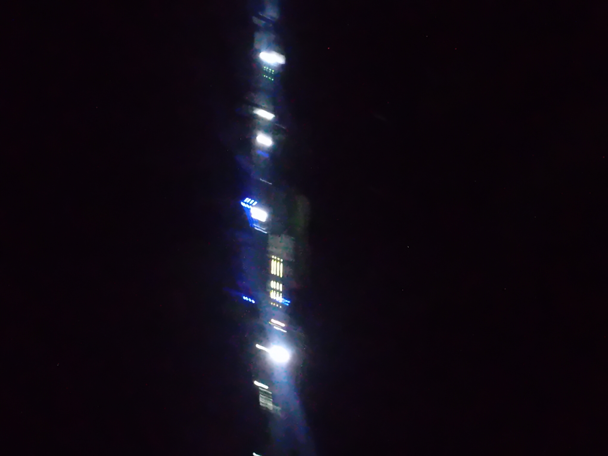 The image is a dark, blurry photograph taken at night. A horizontal band of lights is visible across the middle of the frame. These lights appear to be artificial and are elongated due to motion blur. Some lights are white and bright, while others are blue. There are also some horizontal rectangular patterns of light, possibly indicating windows or illuminated signs on a structure. The background is entirely black, suggesting a very dark environment with minimal ambient light. The image is heavily degraded by motion blur, making it difficult to discern specific details or identify the exact nature of the lights or the location. There are no discernible people, actions, or interactions.