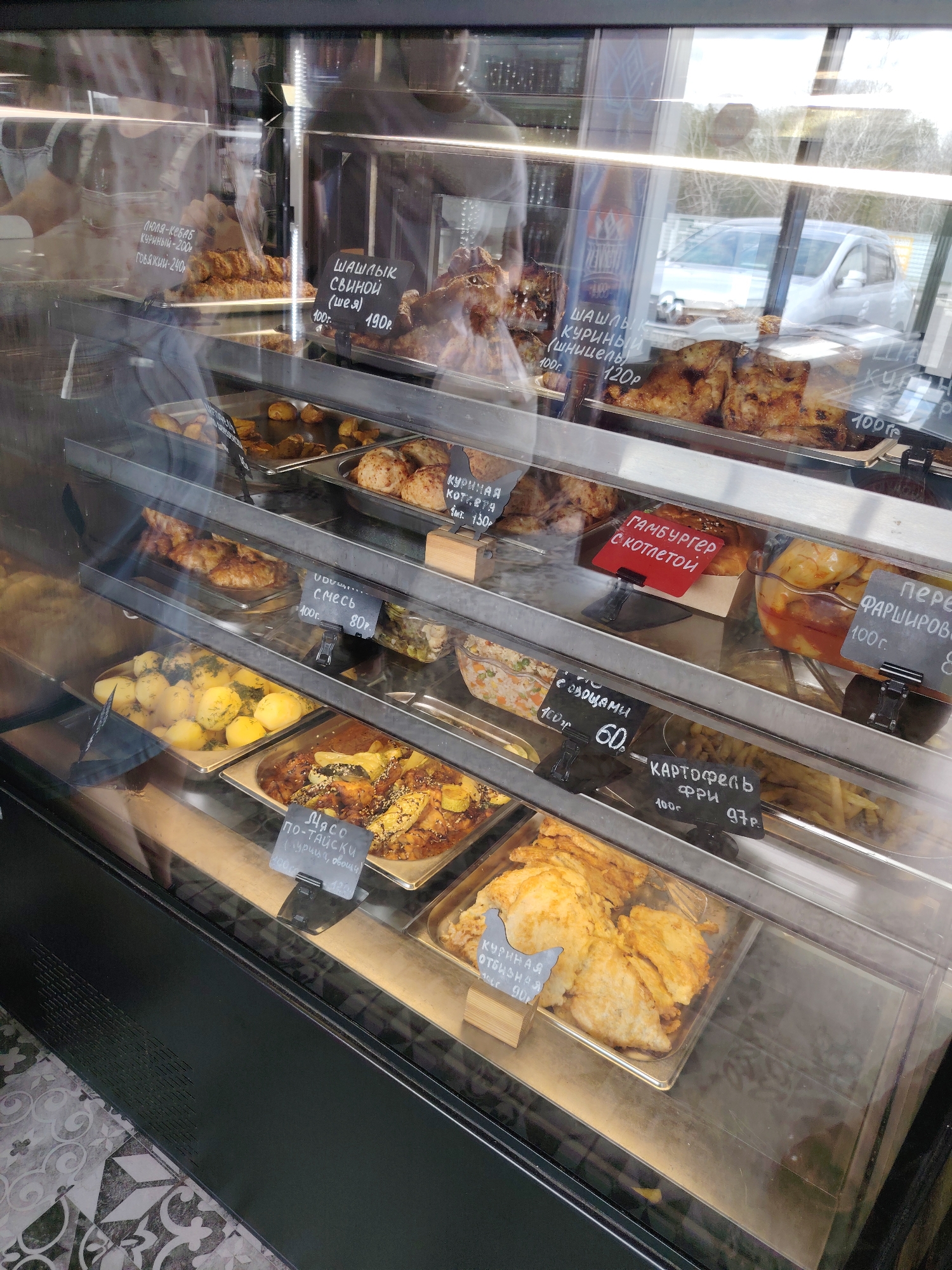 The image shows a refrigerated display case filled with various food items, likely in a cafeteria or food service establishment in Kagal'nitskaya, Russia. The display case is made of glass and metal, with multiple shelves holding trays of food.
On the top shelf, several items are visible, including what appears to be grilled meat or chicken. A sign in Russian indicates "шашлык свиной (шея)" (pork shashlik, neck) with a price of 190 rubles. Another sign says "шашлык куриный" (chicken shashlik) priced at 120 rubles.
The middle shelves contain more food, including what looks like fried chicken cutlets, potato dishes, and possibly some kind of salad or vegetable medley. Signs in Russian denote prices and descriptions such as "куриная котлета" (chicken cutlet) for 130 rubles, "смесь" (mix) for 80 rubles, and "гамбургер с котлетой" (burger with cutlet) which is a bright red sign. Another sign indicates "перец фаршированый" (stuffed pepper) at 80 rubles.
The bottom shelf displays potatoes, a dish labeled "мясо по-тайски (курица, овощи)" (meat in Thai style (chicken, vegetables)) with a price of 120 rubles. French fries are also visible, labeled as "картофель фри" (french fries) for 97 rubles per 100 grams. Another sign points to "куриная отбивная" (chicken cutlet) for 90 rubles.
The overall environment appears to be indoors, with reflections of trees and a vehicle visible through the glass of the display case, suggesting a view of the outside. The flooring below the display case has a patterned design. There are no identifiable people in the frame.