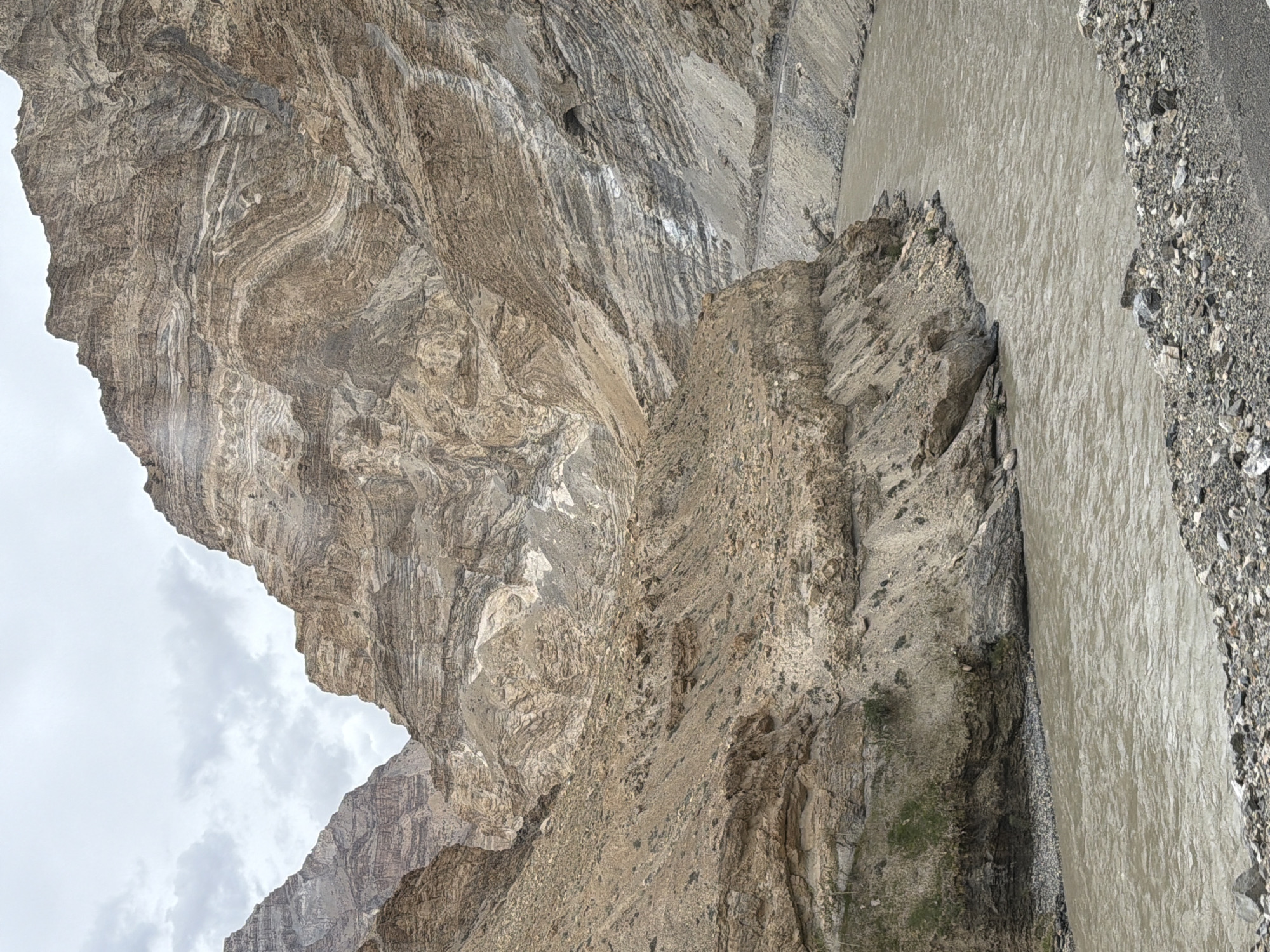 A wide, muddy river flows through a narrow gorge with steep, rocky mountains on either side. The mountains display distinct layers of sediment and rock, indicative of geological folding. The sky is overcast with clouds. The scene appears to be a natural landscape, likely in a mountainous region. There are no people or structures visible in the image. The overall impression is one of rugged natural beauty and geological history.