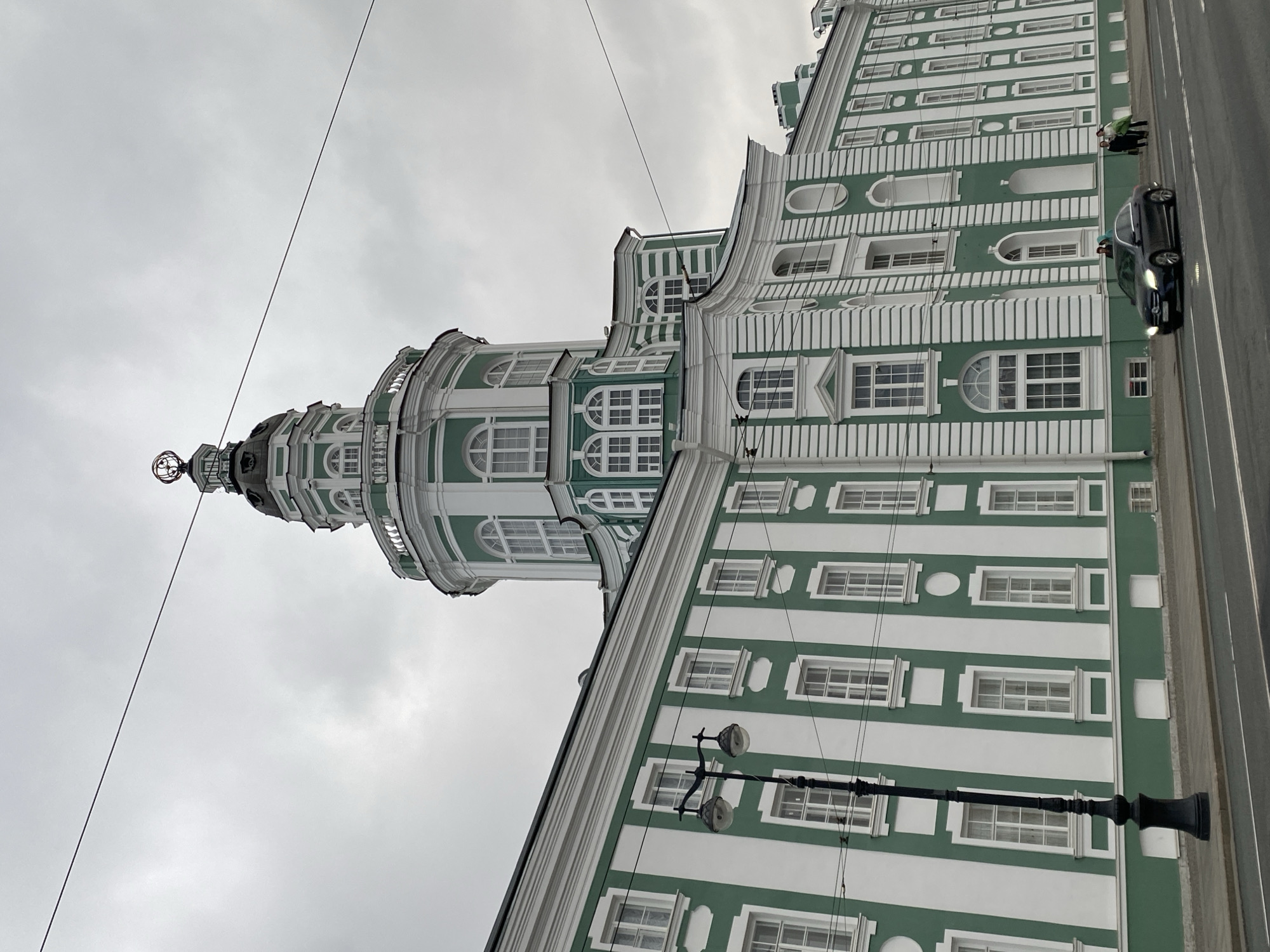 The image shows the Smolny Cathedral in Saint Petersburg, Russia, on an overcast day. The cathedral, painted in white and green, is captured from a low angle, emphasizing its height and architectural details. The distinct onion-shaped dome with a sphere and cross on top is a prominent feature. A black car is parked on the street beside the building. Two people stand on the pavement along the building. Overhead electrical wires traverse the scene. A portion of a black lamppost is visible in the foreground, adding to the urban setting.