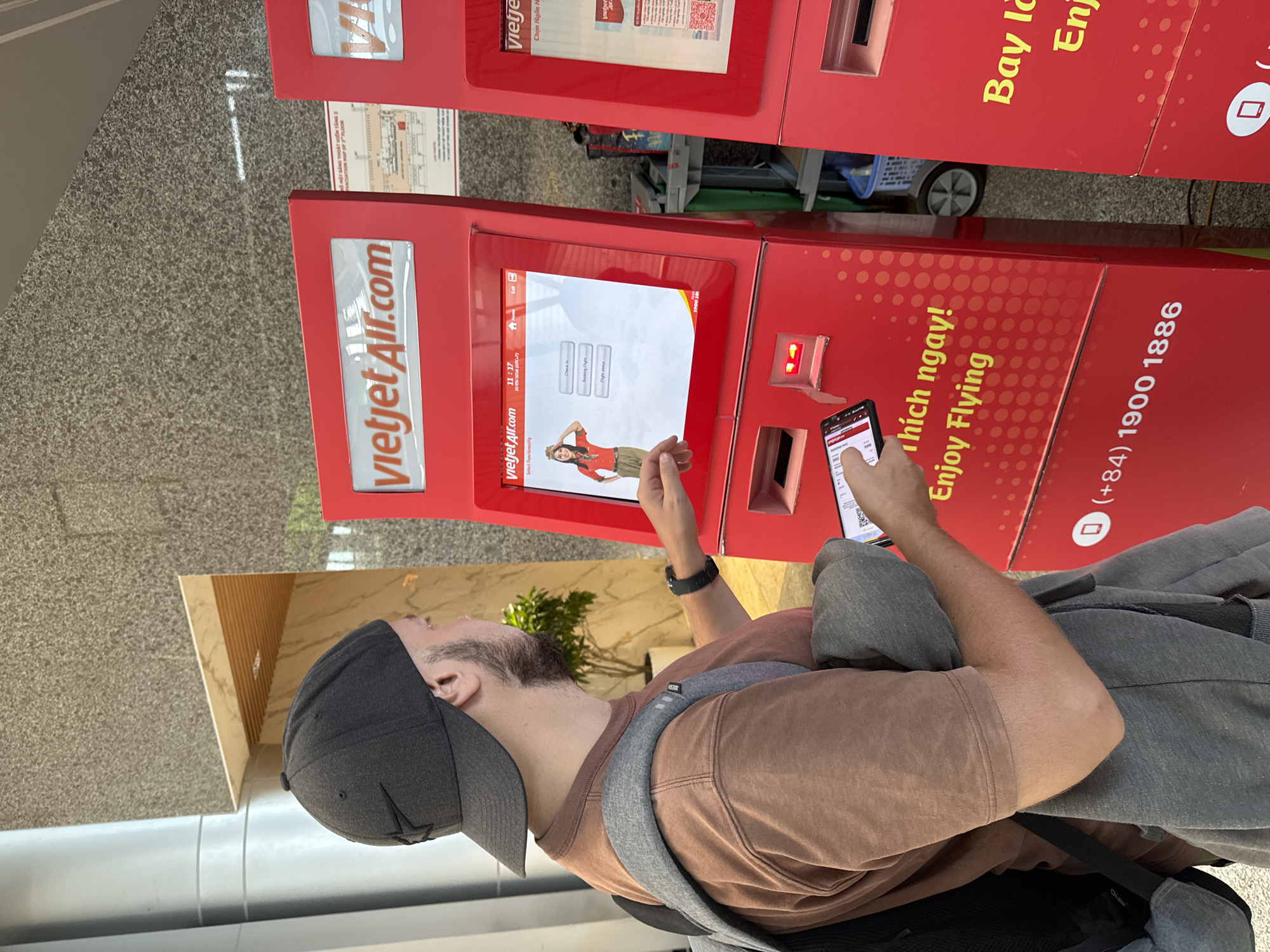 A man wearing a brown shirt and a dark gray baseball cap is standing in what appears to be an airport terminal in Đà Nẵng, Vietnam. He is using a Vietjet Air check-in kiosk. He is also holding and looking at a smartphone in his right hand, while his left hand is on the kiosk screen. The kiosk is red and displays the Vietjet Air logo and the phrase "thích ngay! Enjoy Flying," with Vietnamese text as well. Behind the man, there is another similar kiosk, along with a wall with striped décor. The environment suggests a public area, possibly a terminal at the airport.