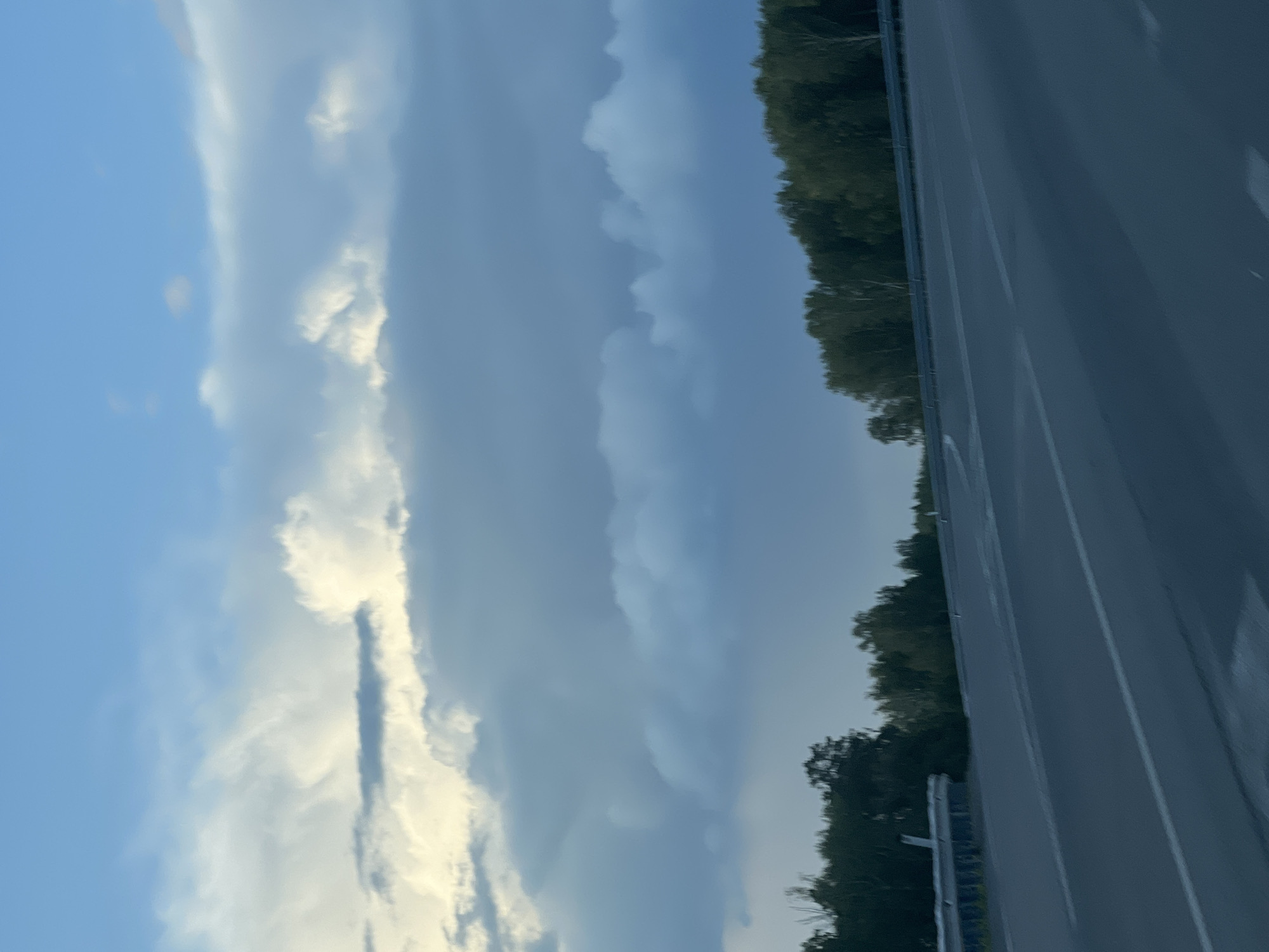 The image shows a highway on a cloudy day, with trees lining the sides of the road. The sky is a mix of blue and gray clouds, with some patches of sunlight breaking through. The highway appears to be a multi-lane road with clear lane markings. There are no people or vehicles visible in the image. The scene suggests a rural or suburban setting outside of a city.