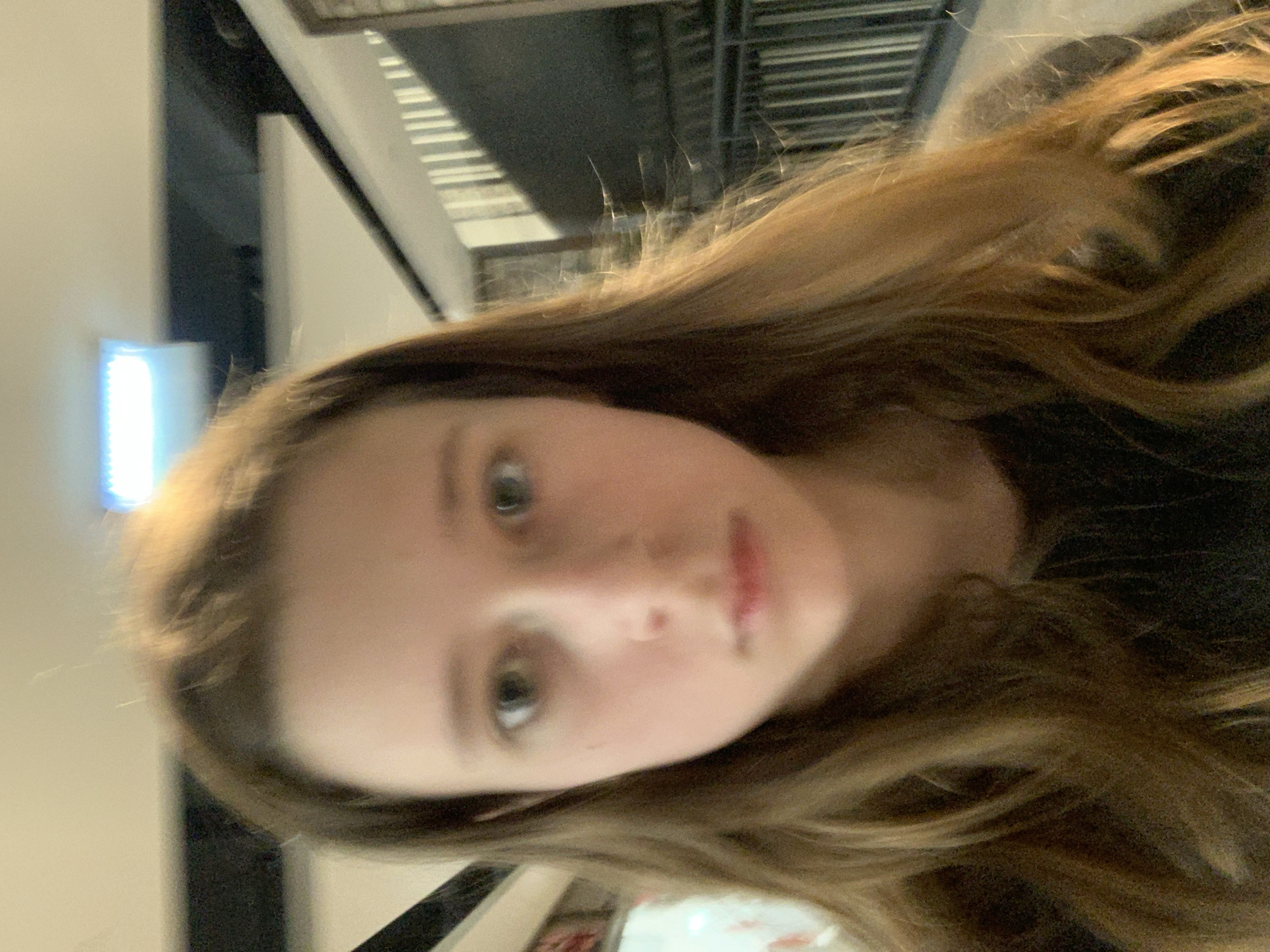 A young woman with light brown hair and fair skin is posing for a selfie indoors. Her eyes are green, and she has a neutral expression. She is wearing a dark-colored top. Behind her, there is a background featuring what appears to be a modern building interior with metal structures and white walls. The lighting is a mix of natural and artificial, with a bright light source visible in the background.
Given the image, there is no verifiable information regarding the location in the city of Belgrade, Serbia.