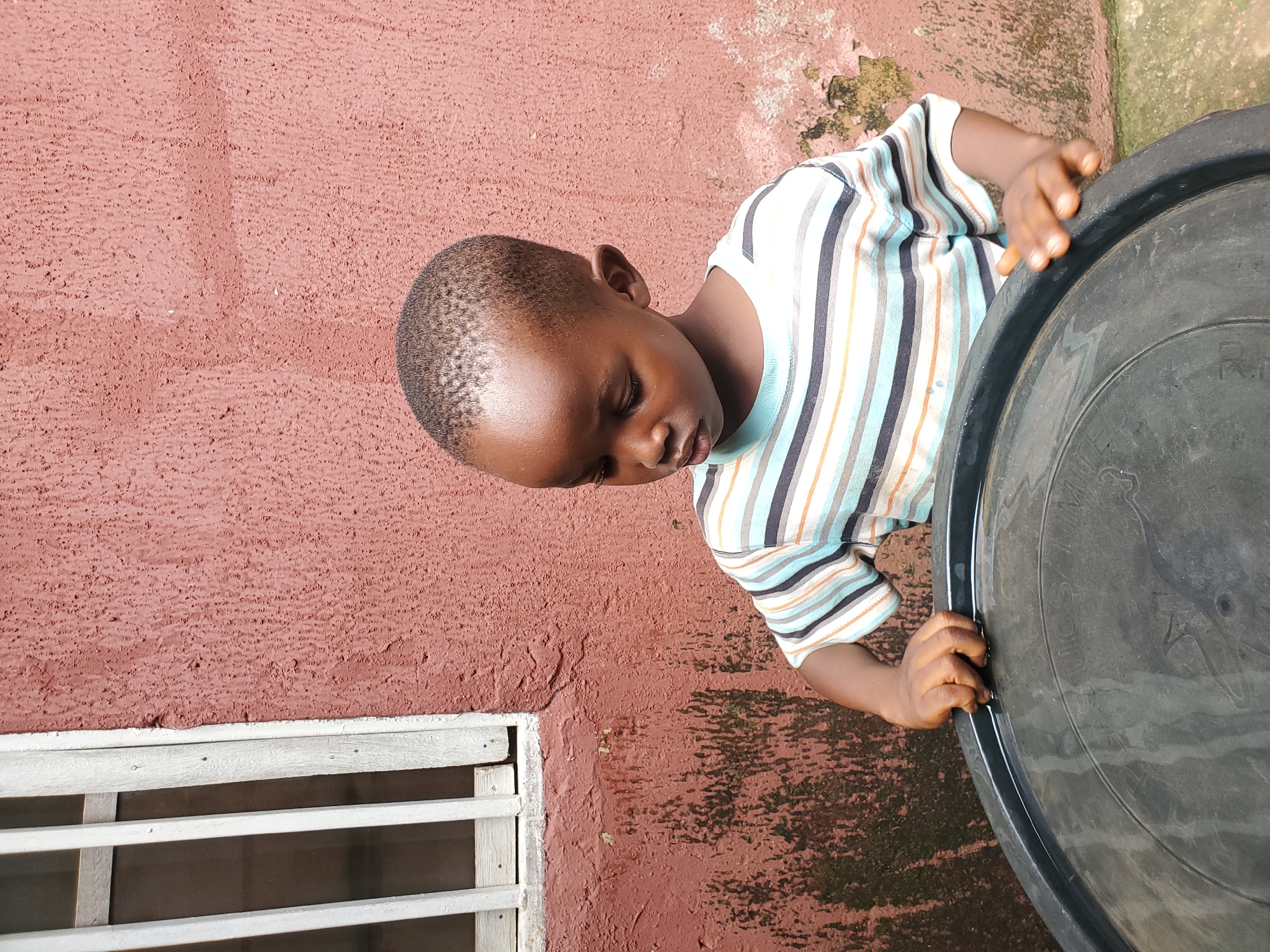 The image shows a young Black child, likely a boy, looking down with closed eyes, appearing contemplative or tired. He is wearing a striped t-shirt with horizontal bands of blue, white, and orange. His hands are resting on the rim of a large, black plastic container, possibly a water tank or barrel. The child is positioned in front of a textured, reddish-brown wall, which appears to be an exterior surface. To the left of the child and wall, there is a white-barred window. The overall setting suggests an outdoor, possibly domestic, environment. The lighting indicates daytime, but the time of day is not precisely discernible. There is no discernible text in the image. The scene appears calm and quiet, with no clear indication of a specific event or strong emotion other than the child's subdued posture. The context provided places the location in Jos, Nigeria.