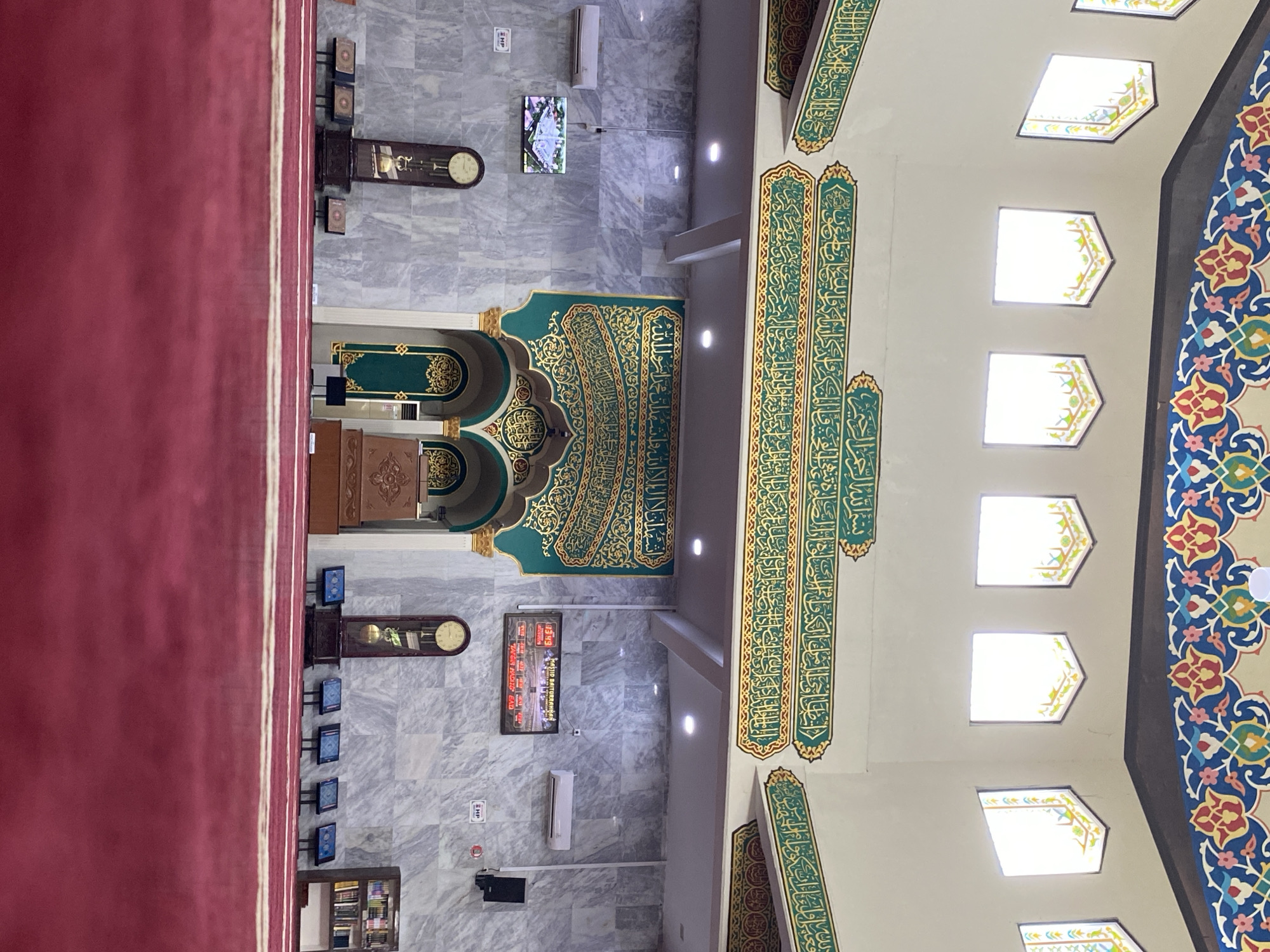 The image shows the interior of a mosque with a red carpeted floor. The walls are adorned with intricate Islamic calligraphy in gold and green. The ceiling features a colorful, ornate mosaic pattern. There are several arched doorways and windows. On the left side, a tall grandfather clock stands next to a small television screen displaying an image. A row of small, dark chairs is visible in front of the clock. On the right side, there is another grandfather clock and a bookshelf filled with religious texts. Several small screens are placed on the floor in front of the chairs. An air conditioning unit is mounted on the wall. The scene appears to be a quiet, solemn space within the mosque, likely in Lhokseumawe, Indonesia, given the presence of the text "MASJID BAITURRAHMAN LHOKSEUMAWE" on one of the screens. There are no people visible in the image.