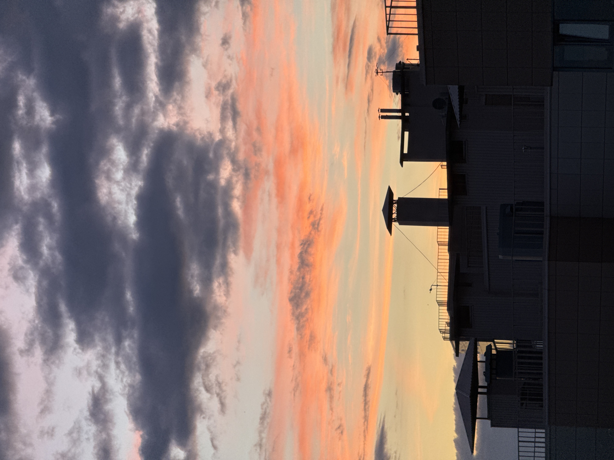 This is a photograph of a city skyline at sunset. The sky is filled with dramatic clouds in shades of pink, orange, and dark gray. Silhouetted against the colorful sky are the rooftops of several buildings, including chimneys and antennae. The buildings appear to be made of concrete and metal, with some featuring balconies or rooftop terraces. The overall impression is one of urban architecture set against a vibrant natural phenomenon. The image does not contain any people or visible actions. The location is identified as Unknown, Russia.