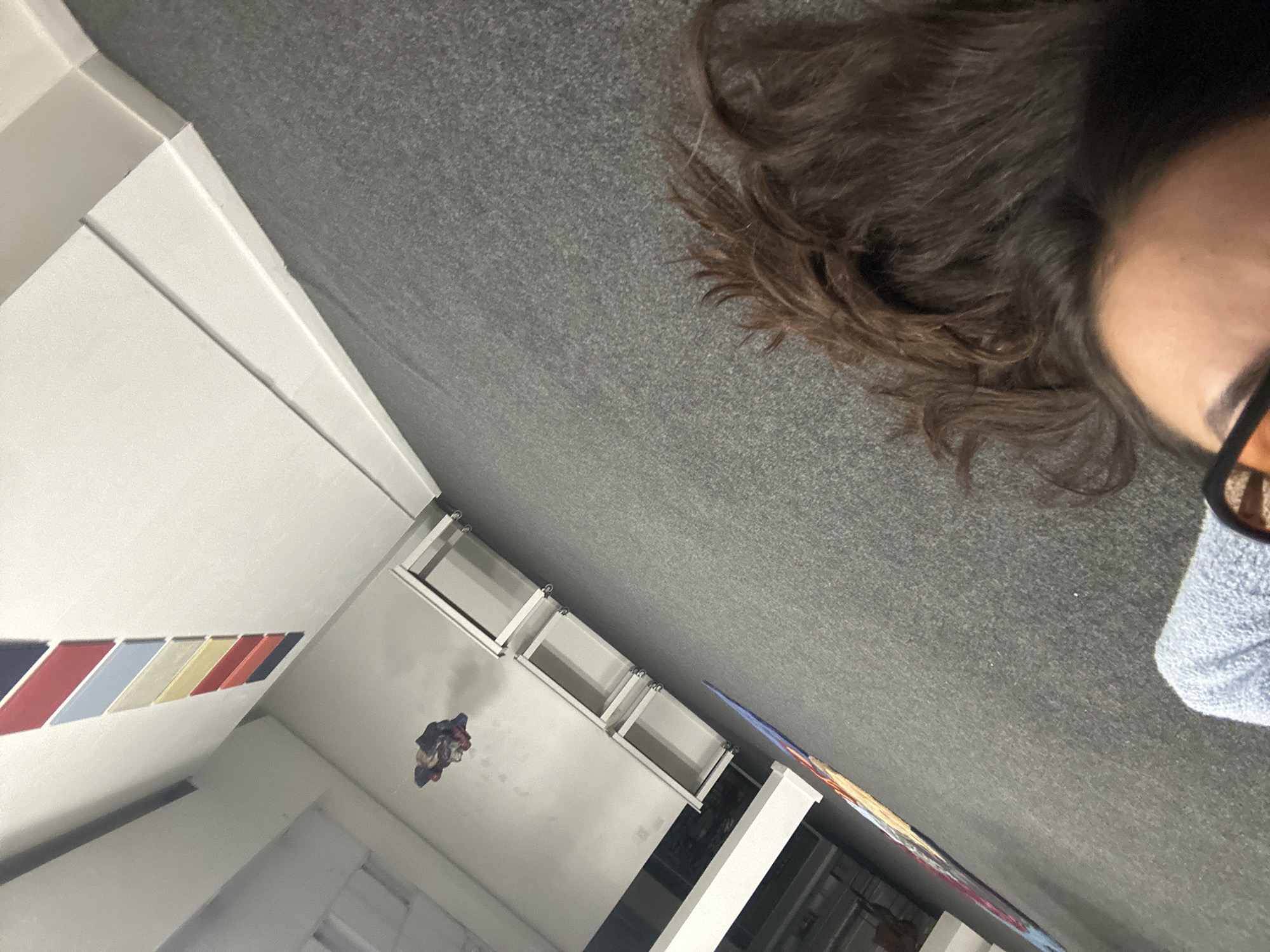 The image presents an indoor scene captured from an unusual, possibly overhead, perspective. In the immediate foreground, on the right side, a portion of a person's head is visible. They have dark brown, wavy hair and wear orange-tinted glasses, with a small section of their forehead and eye visible. The person appears to be resting against or situated above a grey, textured surface, likely carpeting, while wearing light blue, textured clothing.
The background features a predominantly white wall. A notable decorative element on this wall is a horizontal pattern of seven rectangular stripes in distinct colors: dark blue, red, light blue, cream, yellow, red, and dark blue, strongly reminiscent of the BMW M performance brand colors. Below this striped decoration, a series of at least three white, open-faced shelving units or cubbies are mounted on the wall. Further down the wall, a crumpled piece of multi-colored fabric, possibly clothing, is hanging. To the lower right, a colorful, abstract poster or artwork in shades of blue, orange, and white is partially visible.
The overall impression is a casual, personal shot taken from a unique angle within a modern or minimalist interior space. No specific activity is clearly depicted beyond the person's presence. There is no visible text in the image. This scene is situated in Buenos Aires, Argentina.