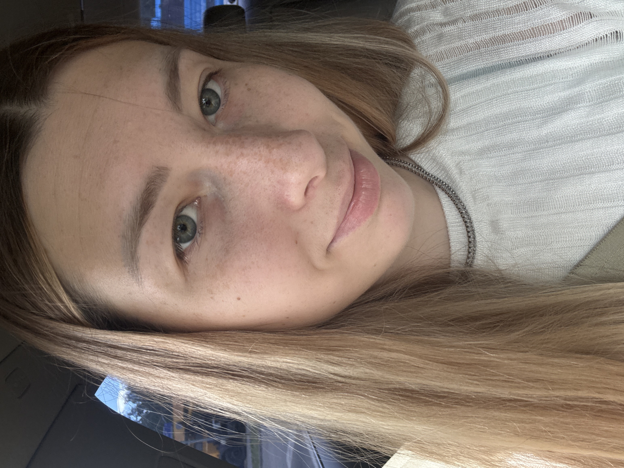 A close-up portrait shows a fair-skinned woman with light brown hair and freckles. She has light green eyes and pink lips. She is wearing a white knitted top with a silver necklace. The background is blurry and suggests an interior, possibly a car. There is no indication of Moscow, Russia, in the image.