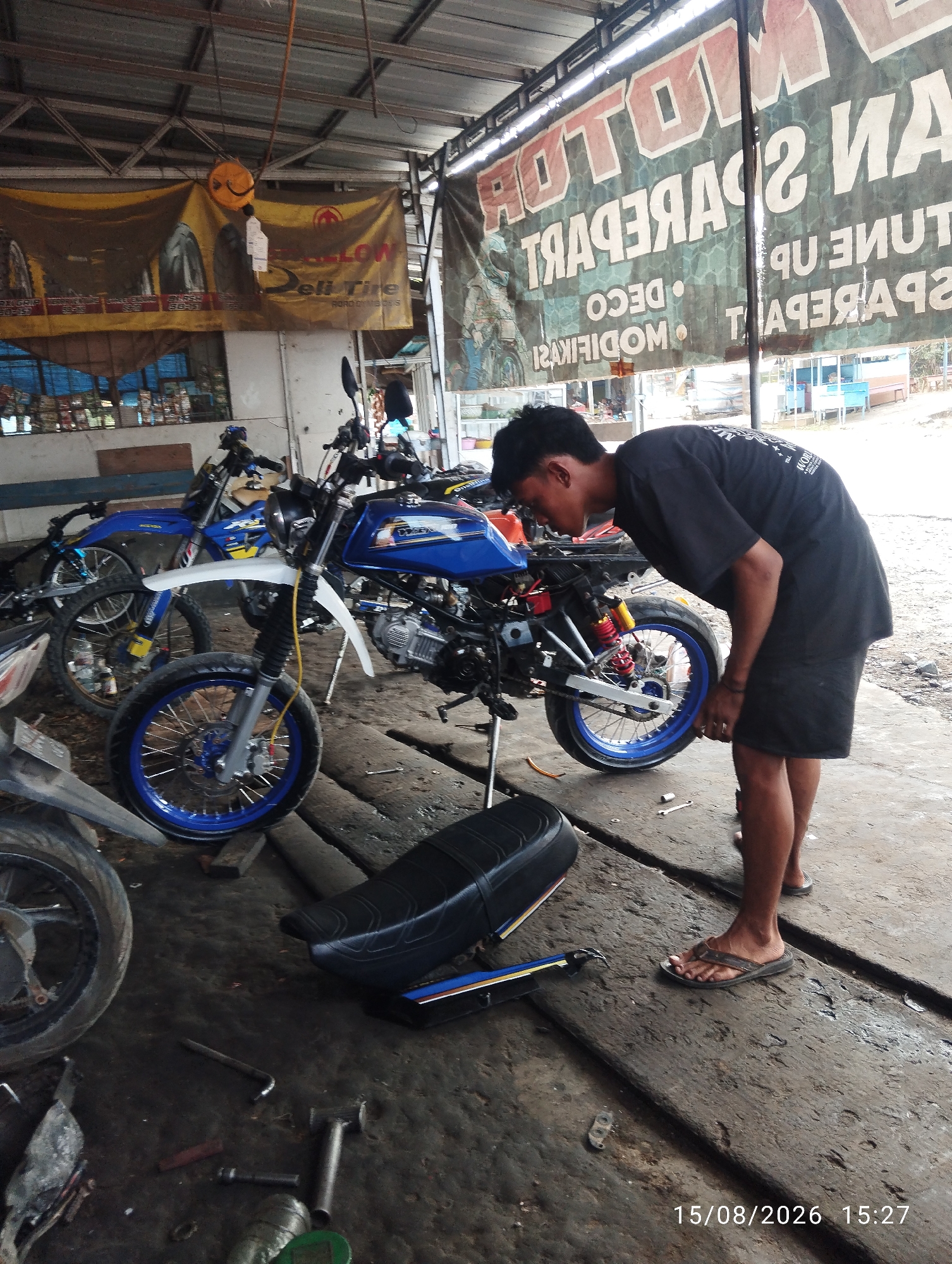 The image depicts a motorcycle repair shop in Tegal Rejo, Indonesia. A young man, wearing a black t-shirt and black shorts, is shown bending over a blue and white motorcycle. Tools, including wrenches and what appears to be a tire lever, are scattered on the ground around the motorcycles. The shop itself appears to be an open-air structure with a corrugated metal roof supported by metal beams. Banners are hung in the background; one appears to advertise tires and the other motor spare parts. The floor is made of concrete and is wet in places, suggesting recent cleaning or a leaky roof. Several other motorcycles are visible in the background. The timestamp on the image reads "15/08/2026 15:27".