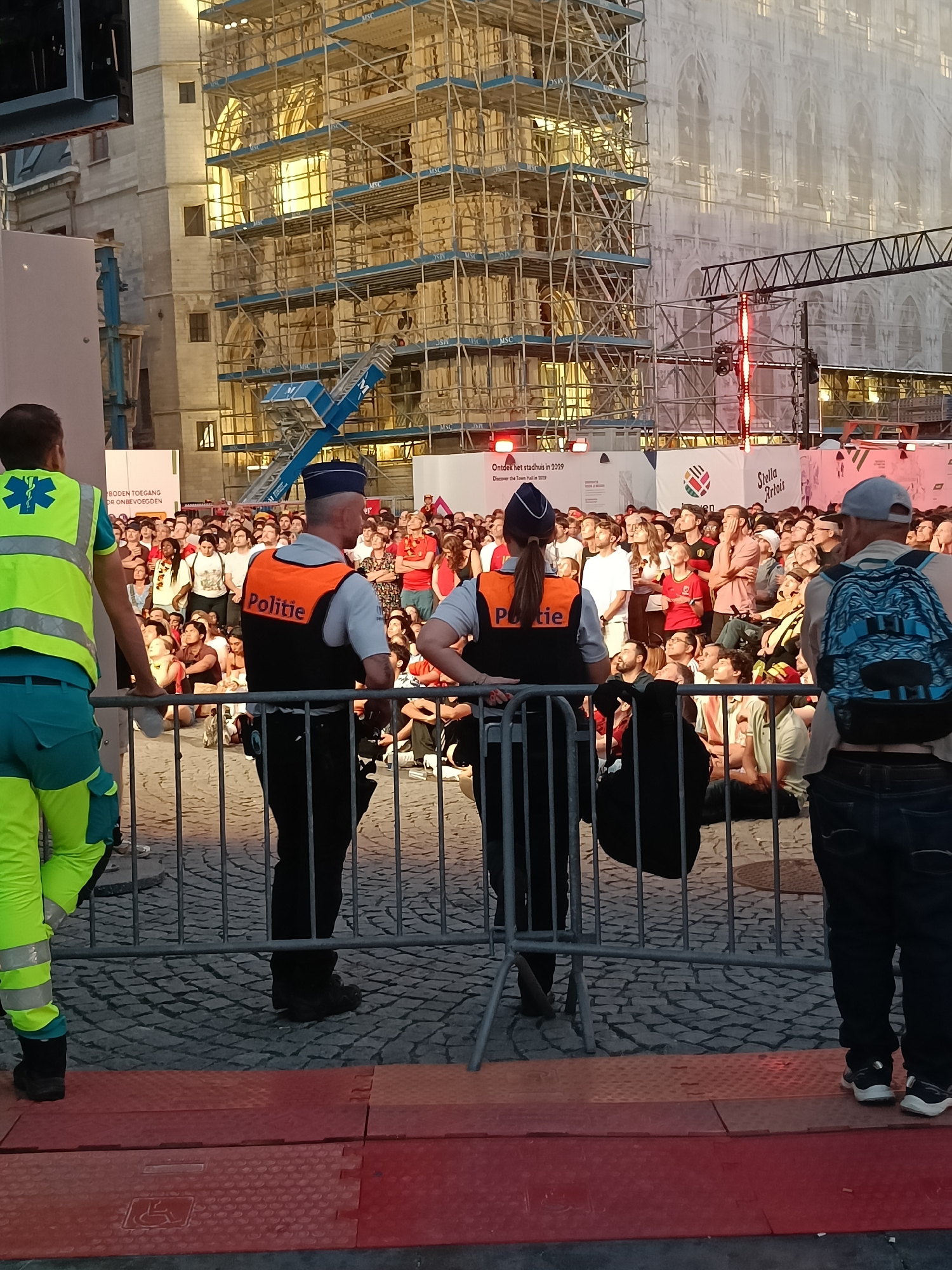 The image depicts a scene in front of the Stadhuis (Town Hall) in Leuven, Belgium, which is undergoing renovation, as indicated by the scaffolding and sheeting covering the building. In the foreground, two police officers and an emergency responder stand behind a metal barrier, facing a crowd of spectators. The police officers wear orange vests with "Politie" written on them. The emergency responder wears a bright yellow-green vest with a blue medical symbol. The crowd appears to be watching an event or performance, with many people looking upwards. The ground is paved with cobblestones. The time of day appears to be late afternoon or early evening, with the lighting suggesting a warm, possibly sunset, glow.
