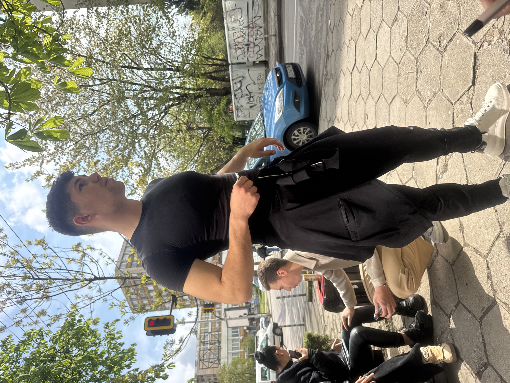 An outdoor urban scene is captured from a low-angle perspective, depicting several individuals on a sunny day. In the foreground, a young man with short dark hair, wearing a black short-sleeved top, black cargo-style pants, and white high-top sneakers, stands with a black bag slung over his shoulder. He is looking upwards and to the right, towards the bright sky and overhead tree canopy.
Behind him and to the right, other people are visible. One individual, wearing a light-colored jacket and tan trousers, is bending down. Further back, two people are seated on the ground or a low structure; one has dark hair tied in a bun and is engrossed in a phone, dressed in black.
The ground is paved with distinctive hexagonal stones, with strong sunlight casting clear shadows. In the background, a light blue car, possibly a Ford Focus, is parked on the street. Its license plate partially shows "CA 0...". Behind the car, two large white containers are covered extensively with illegible graffiti. Abundant green trees with new spring leaves fill the upper and left parts of the frame, silhouetted against a bright blue sky with some white clouds. A red traffic light is visible on the left side, partially obscured by foliage. The scene suggests a casual gathering or simply people going about their day in a bustling city environment.