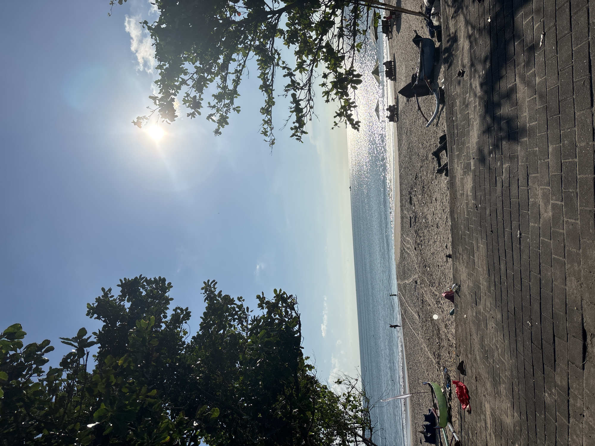 The image shows a sunny beach scene in Kuta, Indonesia. The view is oriented vertically, capturing the sky, ocean, beach, and a paved area. Sunlight glares off the water, creating a sparkling effect. Trees are visible in the foreground, partially obscuring the view. Along the beach, there are lounge chairs, umbrellas, and scattered debris. A few people are visible walking on the beach. The paved area runs parallel to the beach and shows signs of wear and tear.