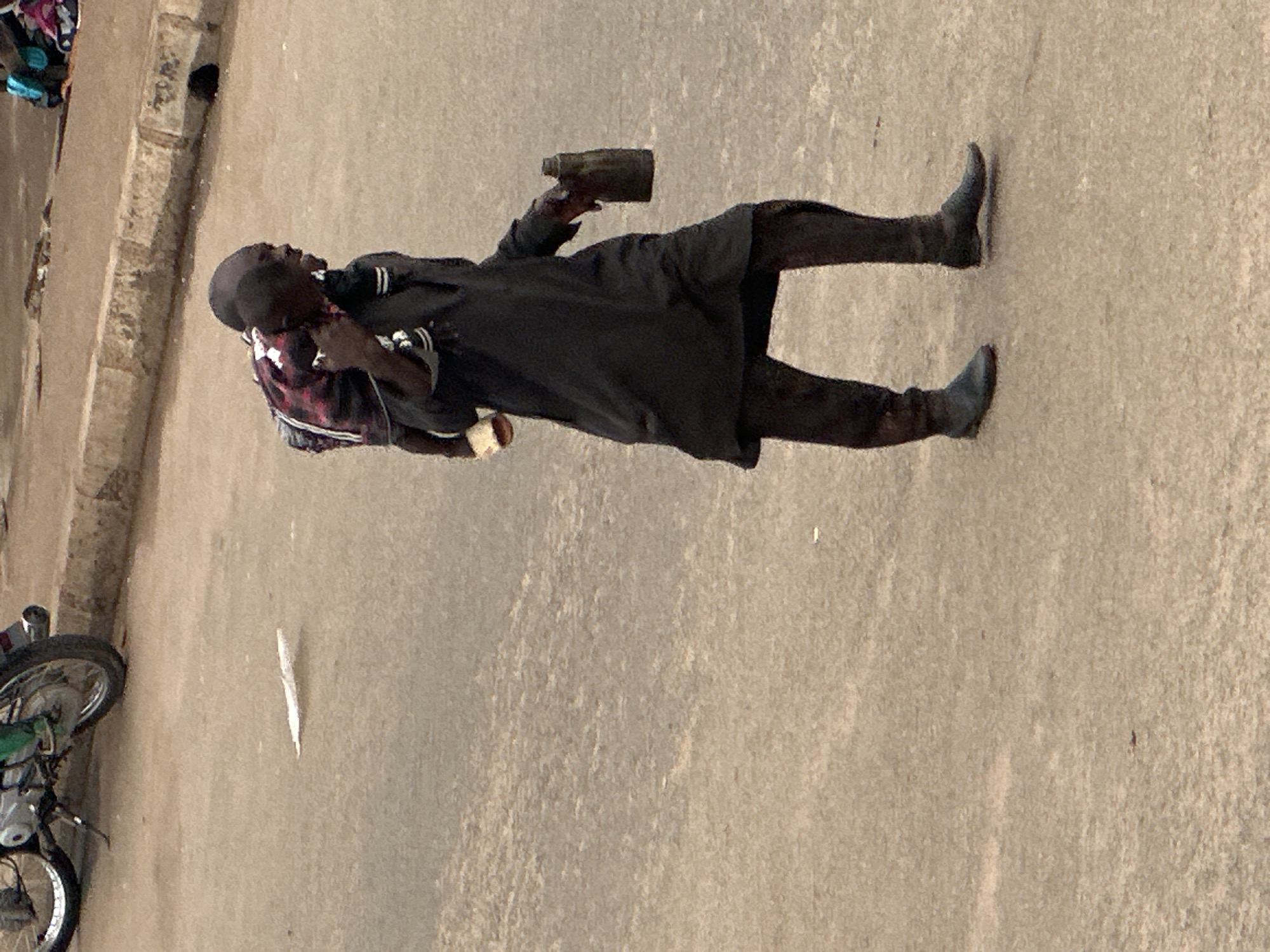 A man carries a child on his shoulders and walks across a paved road in Bukuru, Nigeria. The man is wearing a dark, long-sleeved garment and dark pants. He is also wearing dark boots. The child is wearing a red and black patterned shirt. The man is holding a dark-colored bottle in his right hand. In the background, a motorcycle is parked on the side of the road. The road is made of asphalt and appears to be in a state of disrepair. There is a concrete curb running alongside the road.