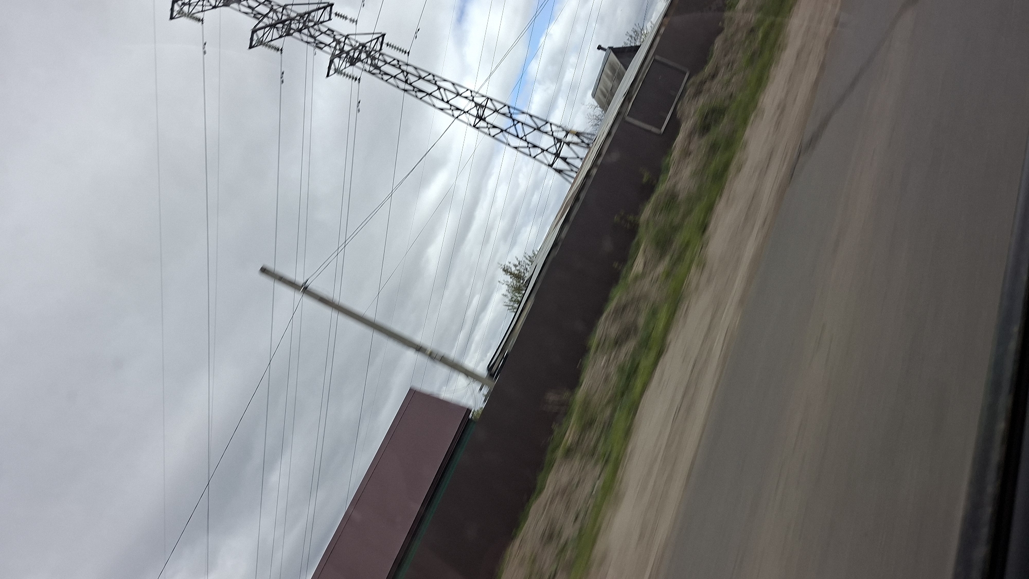 The image depicts an outdoor scene, apparently taken from a moving vehicle on a road. The sky is overcast with dominant grey clouds, with some visible patches of blue. A large electrical transmission tower is prominently featured, with numerous power lines stretching across the sky.
Adjacent to the road is a fence, which is a combination of solid segments and grassy areas. There is a building visible behind the fence, with some windows.
The location is identified as Chaikovsky, Russia.