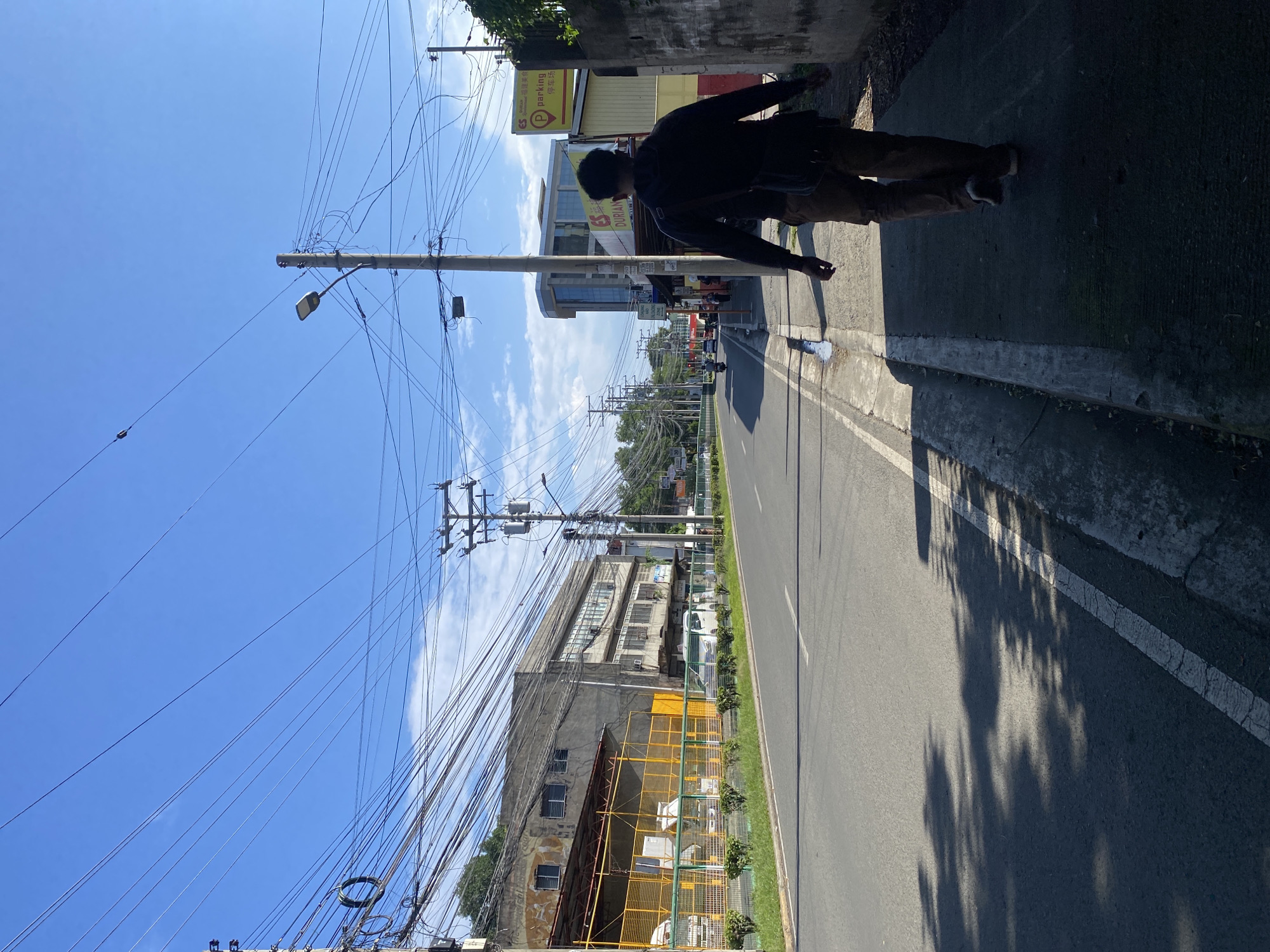 This image depicts a street scene in Davao City, Philippines. A man in a dark hooded jacket and brown pants walks away from the camera along a sidewalk. The sidewalk is next to a two-lane asphalt road, with a white line marking the edge of the road and another line in the middle of the road. On the left side of the road, there is a building with a yellow fence and some trees. On the right side of the road, there are more buildings, including one with a "Parking" sign. The sky is blue with some scattered clouds, and there are numerous electrical wires overhead, strung between utility poles. The lighting suggests it is daytime, likely morning or afternoon, with shadows cast by trees and possibly buildings on the road. The environment appears to be an urban or suburban area with commercial and residential structures lining the street.