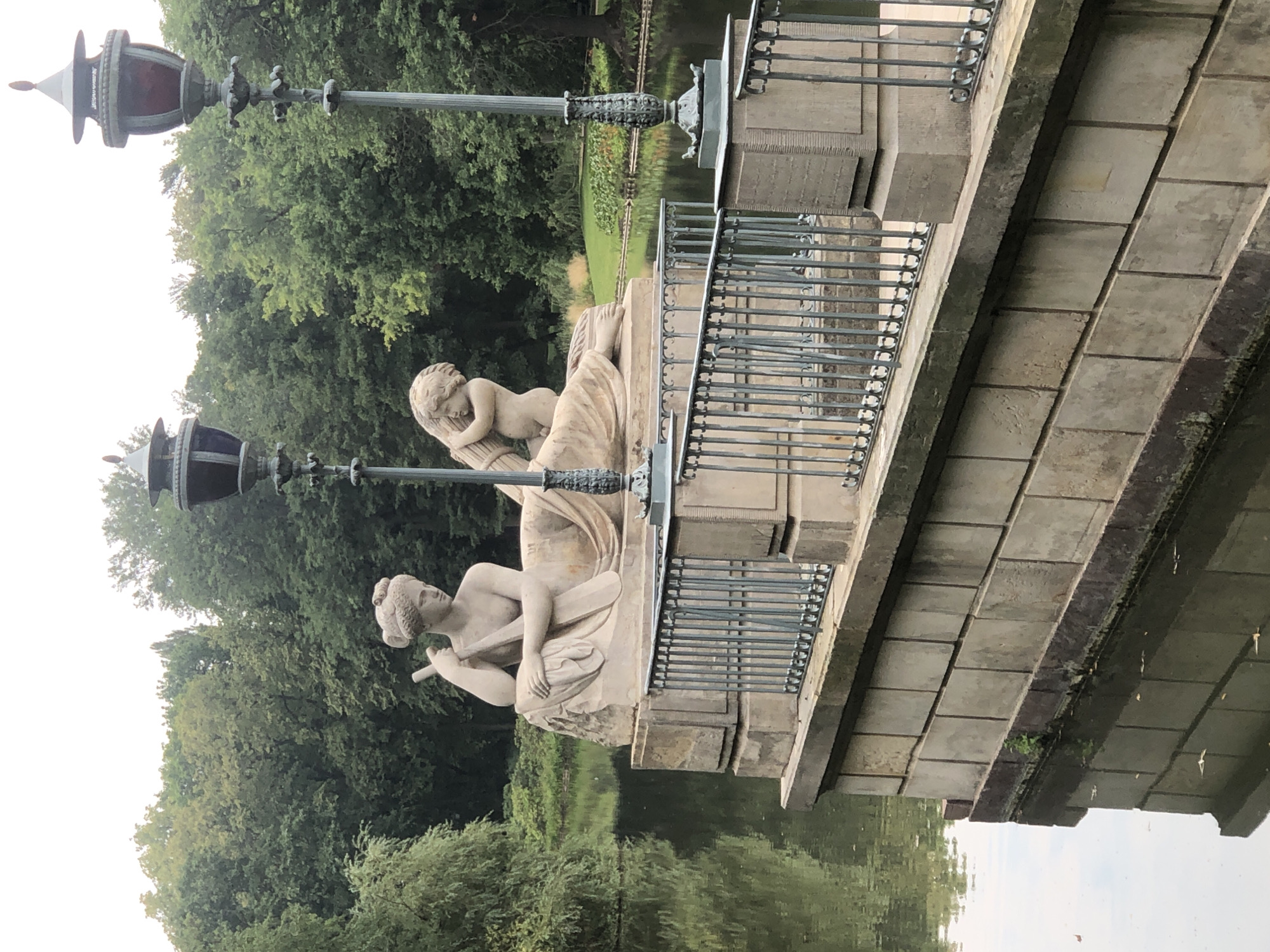 The image depicts a picturesque scene in the Royal Baths Park (Łazienki Królewskie) in Warsaw, Poland. In the foreground, a stone balustrade borders a body of water, reflecting the surrounding lush greenery and overcast sky. Two ornate lampposts, one on each side of the scene, stand tall against the backdrop of dense trees. Atop a stone pedestal, a classical sculpture features a reclining female figure holding what appears to be a staff, and beside her, a cherubic child is embracing a bundle of wheat. The statue, carved from light-colored stone, is detailed and weathered, suggesting its age and exposure to the elements. The overall ambiance is serene and historical, evoking a sense of timeless beauty.