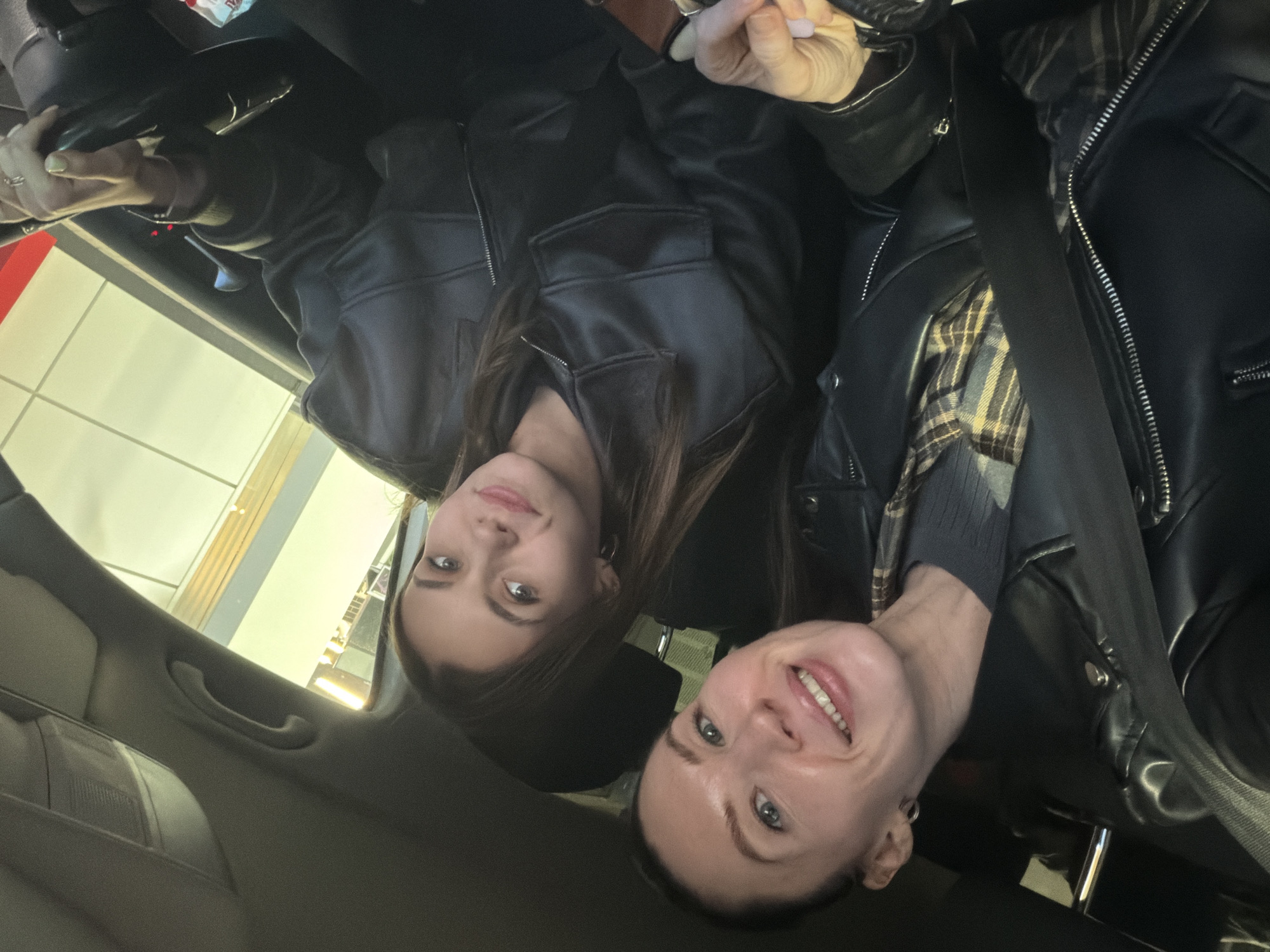 Two women are in a car, likely taking a selfie. One woman has longer brown hair and is wearing a black leather jacket. The other woman has shorter hair and is wearing a black leather jacket over a plaid shirt. They are both smiling. The location is likely in Perm, Russia, based on the available information. The car's interior is visible, including the dashboard and passenger seat. The exterior shows a glimpse of a building and lighting.