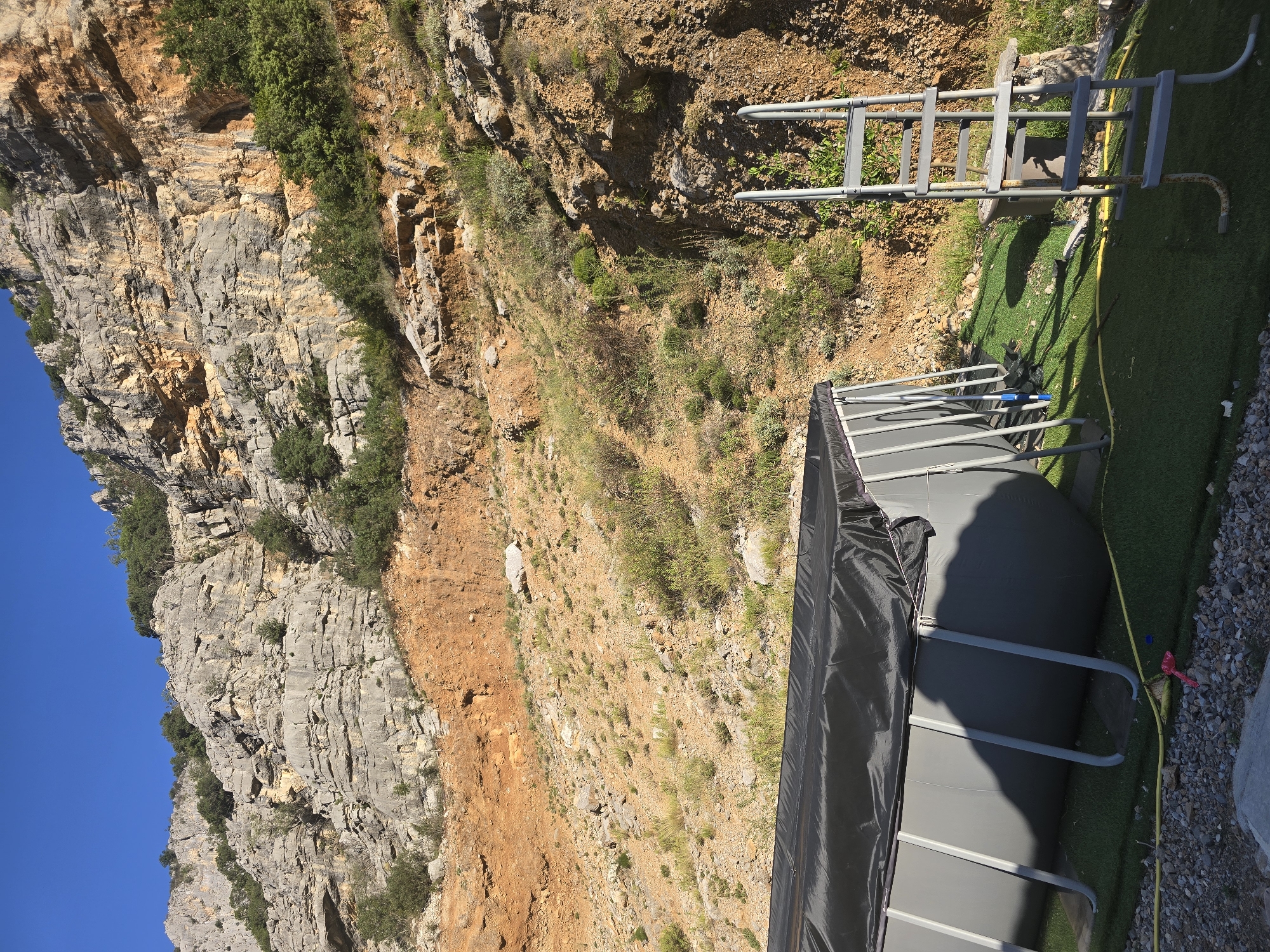 An outdoor above-ground swimming pool, covered with a black tarp, sits on artificial turf next to a rocky, arid hillside under a clear blue sky. A metal ladder leans against the pool, and a yellow hose is visible on the ground. The hillside is characterized by layered rock formations, sparse vegetation, and patches of reddish-brown soil. The scene appears to be in a rural or semi-rural setting, possibly a backyard or recreational area adjacent to a natural landscape.
