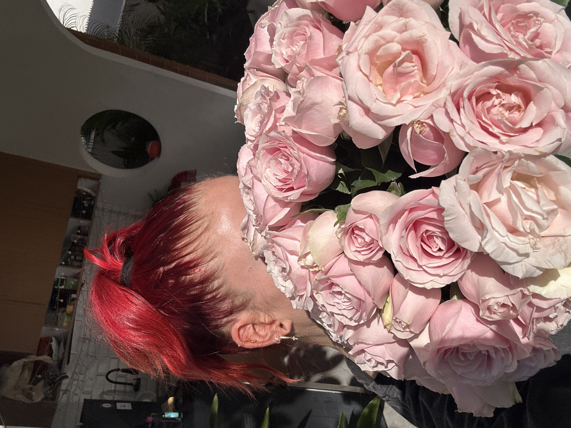The image shows a close-up of a woman with bright red hair pulled back in a ponytail, holding a large bouquet of pale pink roses. Her ear, adorned with multiple piercings and dangling earrings, is visible. The background suggests an indoor setting with a kitchen area visible to the left, featuring white brick tiles and shelves stocked with items. To the right, an arched architectural feature leads to a circular window through which some greenery and shadows are seen. The lighting indicates it is daytime, with strong sunlight casting shadows. The scene does not provide any specific clues about its location in North Kuta, Indonesia.