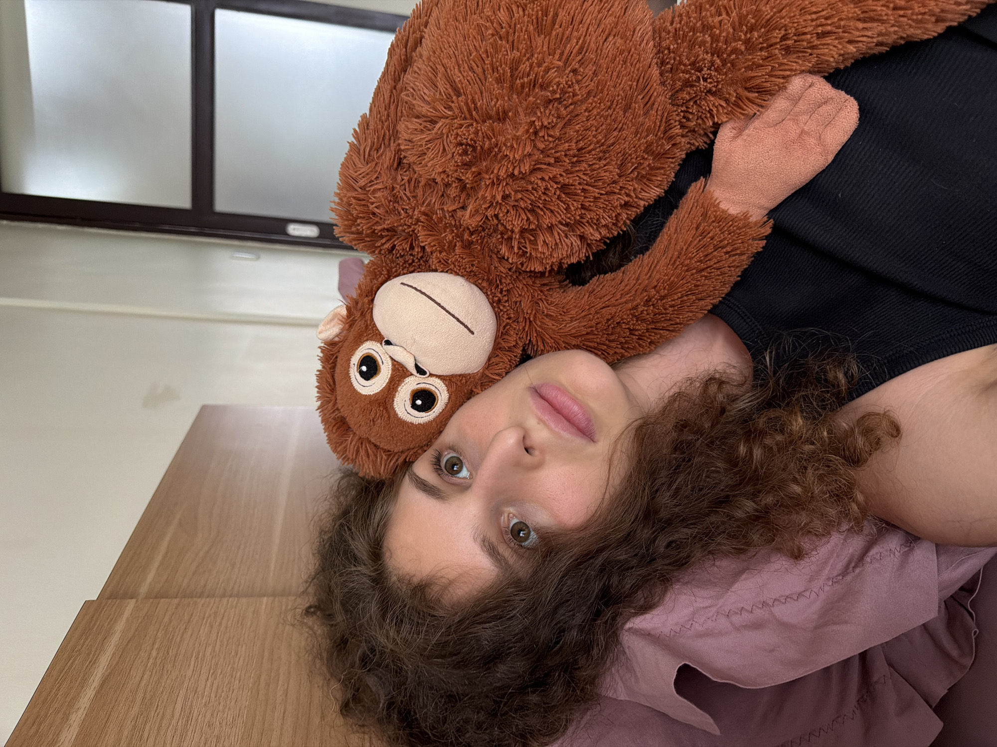 A young woman with curly brown hair lies in bed with a stuffed orangutan. She is on her side, with her head resting on a mauve pillow. The orangutan is brown and fluffy, with large eyes and a smiling mouth. It is positioned so that its arm is around the woman's neck and its head is next to hers. The woman is wearing a black tank top. Behind her, a wooden headboard and a sliding glass door are visible. The scene appears to be in a bedroom in North Kuta, Indonesia, suggested by the tropical climate implied by the presence of a sliding glass door and the likelihood of such accommodations in that region.