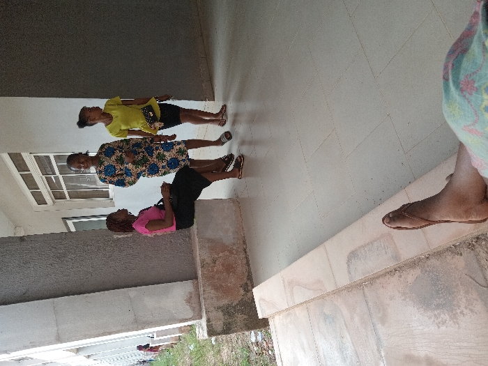 The image captures a daylight scene within a covered walkway or corridor of a building in OGBOKO, Nigeria. In the immediate foreground, the right lower leg and foot of an individual are visible, resting on a beige tiled surface. The foot wears a brown thong sandal and the leg is partially obscured by light-colored, patterned fabric.
In the mid-ground, three adult females are present. On the left, a woman is seated on a low, weathered concrete ledge. She wears a pink short-sleeved top and a black skirt, holding a dark handbag on her lap, and is looking towards the right. Standing in the center, a second woman wears a patterned dress with blue and green motifs on a light background, smiling towards the general direction of the camera. She wears dark open-toed sandals. To her right, a third woman stands, facing right, wearing a bright yellow short-sleeved top, a dark skirt, and a small black handbag with silver-colored accents across her body. Her hair is pulled up.
The environment consists of a building with a light-colored facade, visible windows with white frames, and concrete pillars supporting the walkway structure. The floor is covered with large, light-colored tiles. To the left of the walkway, a strip of ground with sparse vegetation and dirt is visible. The scene suggests a casual social interaction among the three standing and seated individuals.
