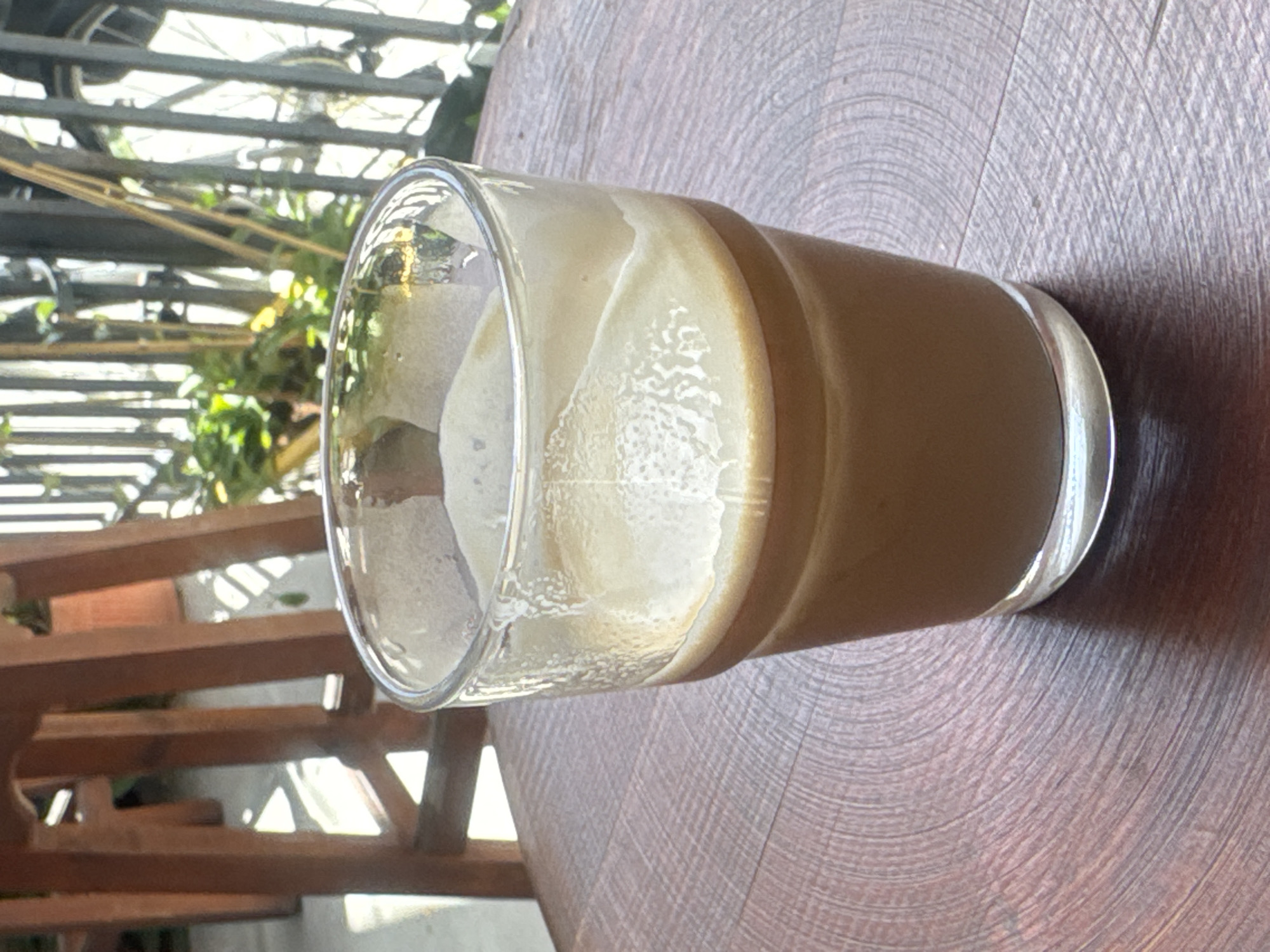 A partially consumed coffee drink in a glass sits on a wooden table. The background shows a balcony railing and plants, suggesting the photo was taken outdoors in an urban environment. Given the prompt, it is implied that this location is Tel-Aviv, Israel.