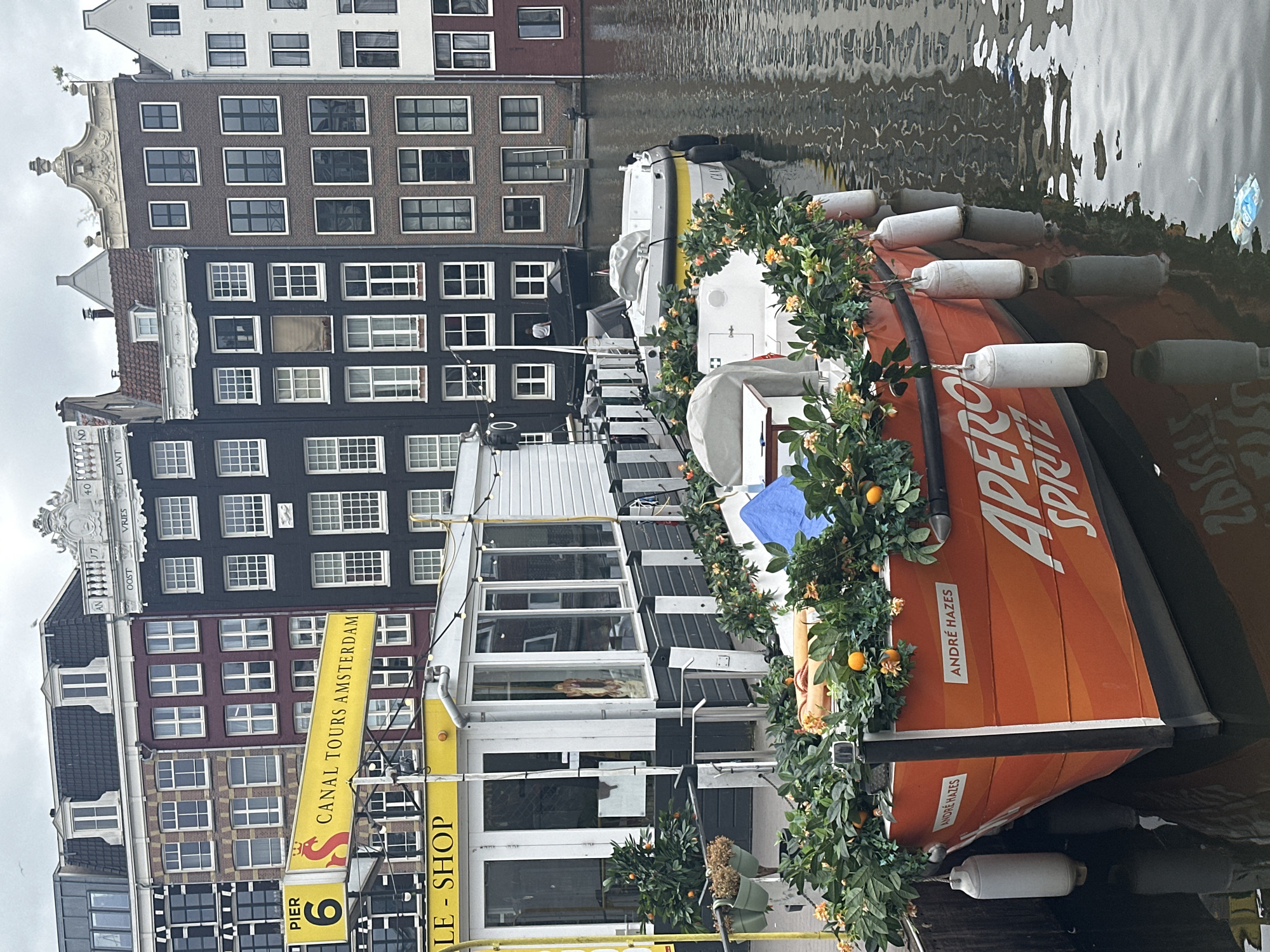 In Amsterdam, Netherlands, an orange boat named "APERO SPRITZ" is docked at Pier 6 of Canal Tours Amsterdam. The boat is adorned with a lush green garland of leaves and small orange decorations, creating a festive atmosphere. Two white buoys are attached to the side of the boat, floating in the calm canal waters. Behind the boat, a white building with large glass windows, likely a ticket office or cafe, is visible. Further in the background, traditional Dutch canal houses with varying facade colors and architectural styles line the waterway under an overcast sky. A few other boats are also present in the canal, and a person can be seen on one of them. The water reflects the surrounding buildings and the cloudy sky.