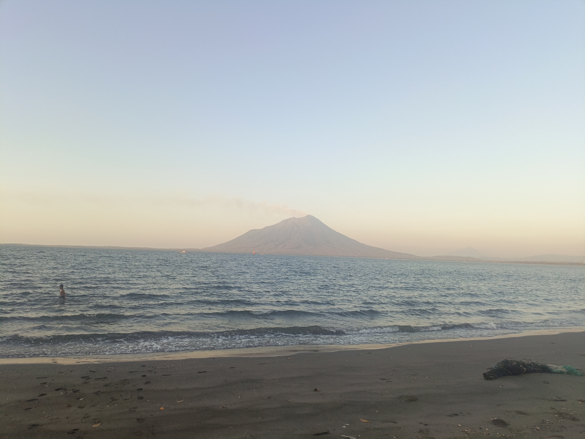 A wide-angle, eye-level shot captures a serene beach scene in Lewoleba, Indonesia, with a large, conical mountain dominating the distant horizon. The sky is a pale gradient of blue and yellow, suggesting either dawn or dusk. The sea, a deep blue with gentle waves, laps against a dark, sandy beach littered with a few fallen leaves. In the foreground, a person stands partially submerged in the water on the left side of the frame. Further out in the sea, a small boat is visible. A plume of smoke rises from the peak of the distant mountain, indicating potential volcanic activity. On the right side of the beach, a bundle of fishing net lies on the sand. The overall atmosphere is tranquil and vast, with the natural elements of sea, sand, and mountain taking center stage.