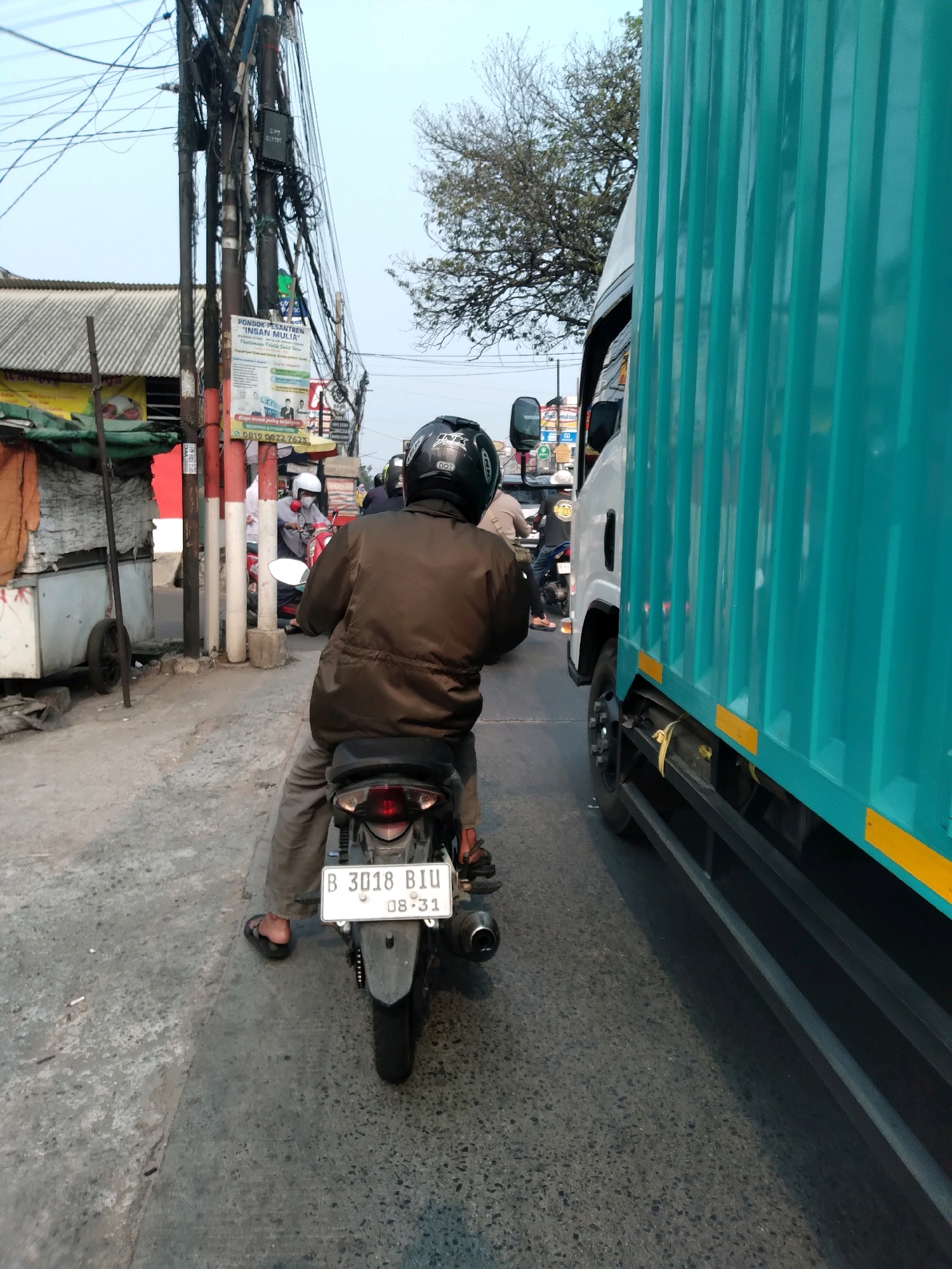 The image depicts a street scene in South Tangerang, Indonesia, likely during the day. In the foreground, a person is riding a motorcycle. They are wearing a brown jacket, grey pants, and a black helmet. The motorcycle has a license plate that reads "B 3018 BIU 08-31". To the right of the motorcycle, a large, turquoise-colored truck is visible, taking up a significant portion of the frame. The truck has a corrugated metal side with yellow reflective tape.
The environment appears to be a busy street with significant electrical wiring overhead, suggesting a developed urban area. There are buildings and other vehicles in the background, including other motorcycles and possibly a smaller car. A street vendor's cart with orange fabric is on the left side of the image. The road surface is asphalt, and there is a slight incline. The sky is a pale blue, indicating daylight. The overall impression is a snapshot of everyday traffic in a bustling Indonesian city.
