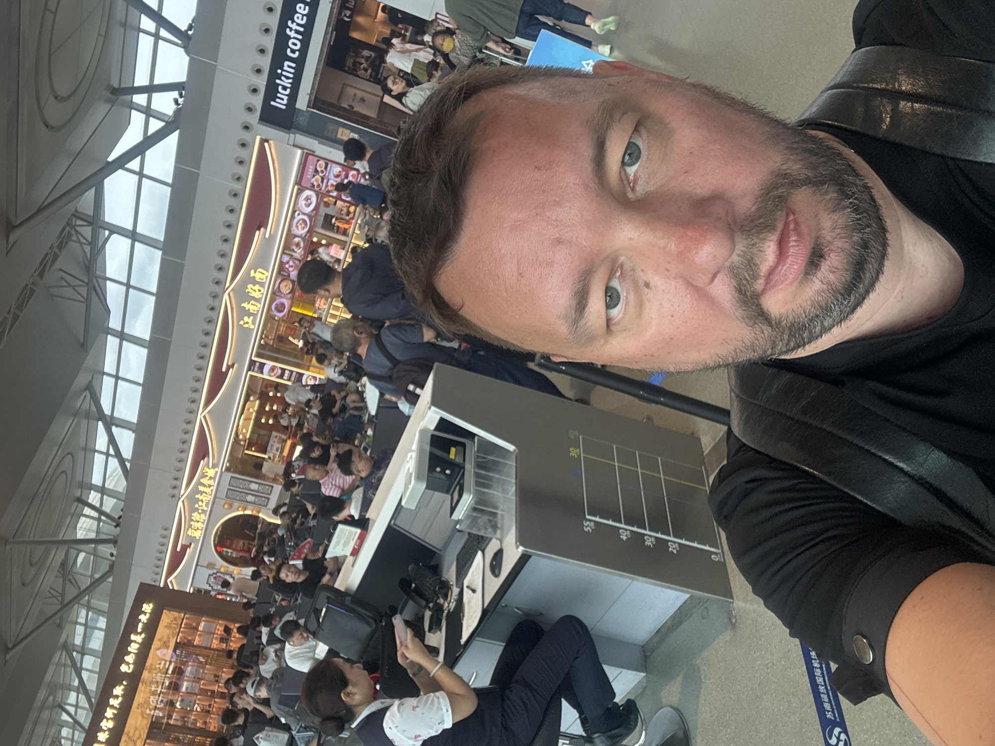 A man takes a selfie in a busy airport terminal. He is positioned in the foreground, with his face filling the bottom half of the frame. He has short, light brown hair, blue eyes, and a beard. He is wearing a black t-shirt and a black leather strap across his shoulder.
In the background, a woman in a dark blue uniform with a white patterned shirt is seated at a counter, appearing to be working. There are phones, a computer monitor, and other equipment on the counter. Behind her, a crowd of people are seated and standing, waiting. Above them, signs in Chinese characters are visible, one advertising "luckin coffee". The terminal has a modern design with a high ceiling and large windows. A blue and white barrier tape with Chinese characters runs along the floor in the foreground. The environment appears to be the interior of an airport in Xinwu District, China, based on the presence of the "luckin coffee" sign and Chinese text on signage.