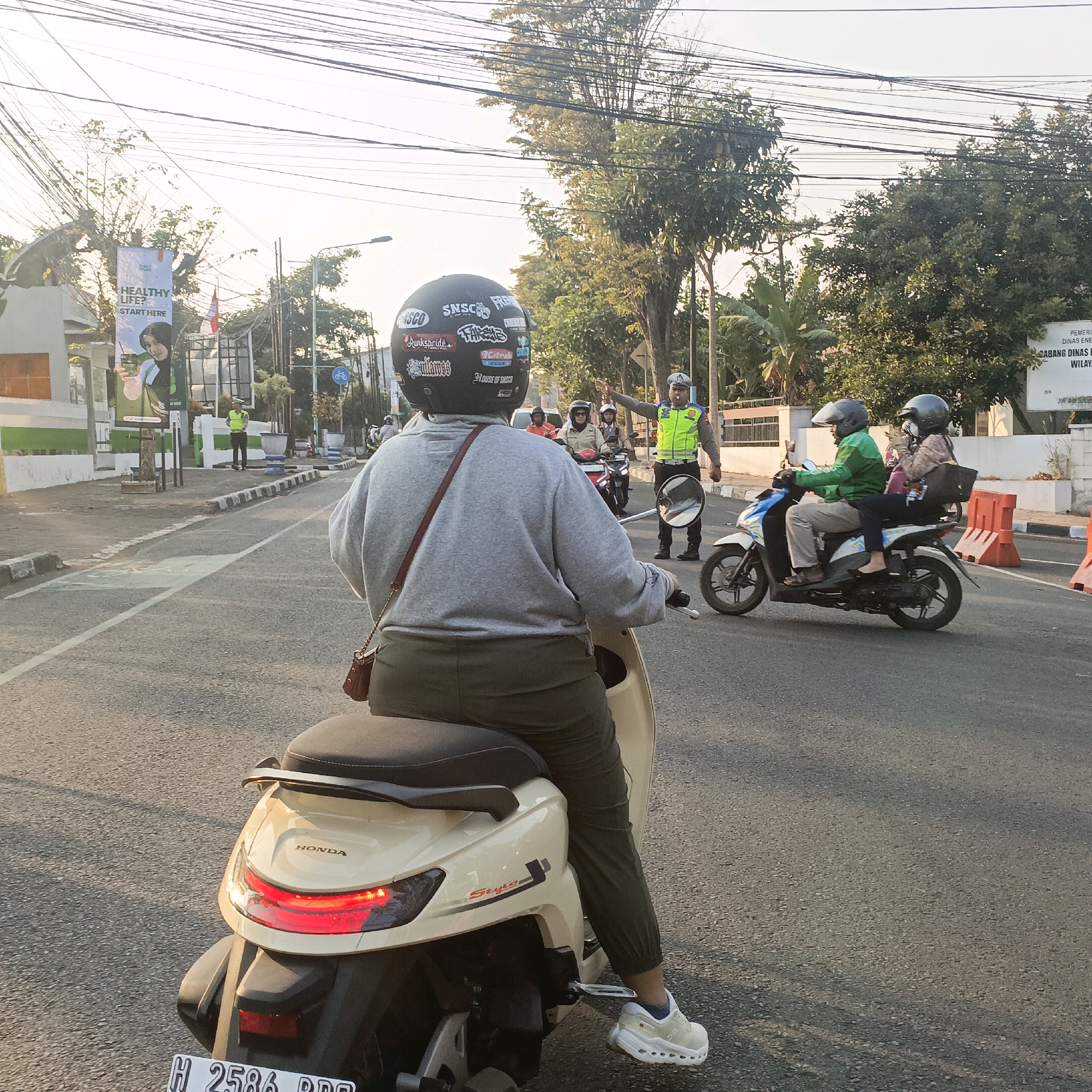 In Salatiga, Indonesia, a person wearing a grey sweater, green pants, and a black helmet with stickers is riding a beige scooter. The scooter's taillight is illuminated. The person is on a paved road with a white line marking the lane. In the background, a police officer in a reflective vest and helmet is directing traffic with their right hand raised. Another scooter carrying two people, one in a green jacket and the other in a patterned shirt and hijab, is to the right of the police officer. There are trees, buildings, streetlights, and power lines visible in the environment. A banner with the text "HEALTHY LIFE? START HERE" is also present. Orange traffic barriers are positioned on the right side of the road.