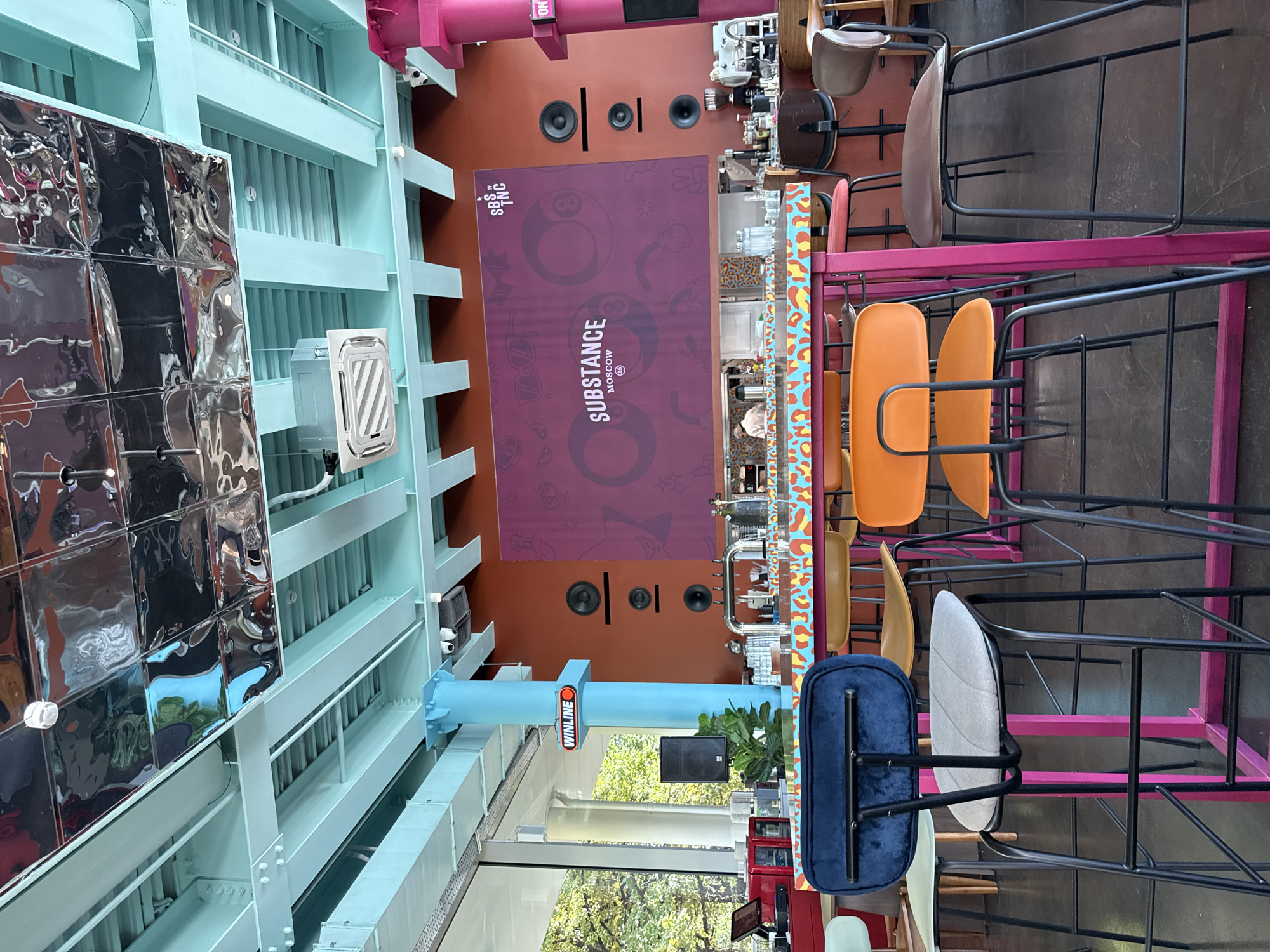 The image shows the interior of a modern cafe or bar, likely in Moscow, Russia, given the "SUBSTANCE MOSCOW" signage. The space features a vibrant and industrial aesthetic with exposed blue beams and a reflective metallic ceiling. Several high bar stools with colorful upholstery are arranged around patterned countertops. In the background, a bar area with coffee machines and bar taps is visible, with a few staff members working. Large graphic artwork adorns the orange wall, featuring bold designs and text. The overall impression is of a trendy and energetic establishment.