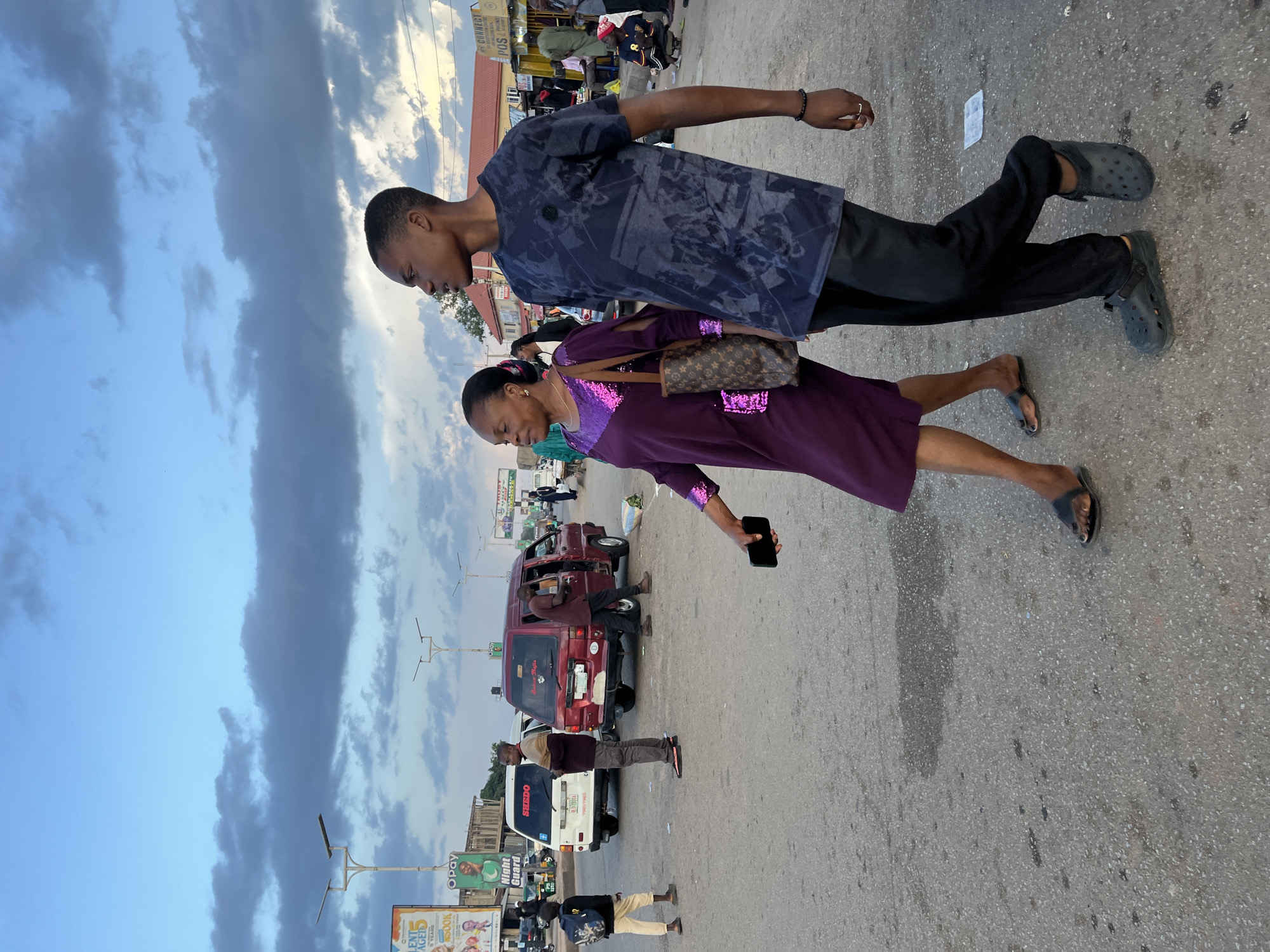 In Gura Top, Nigeria, at dusk, two people walk across a dusty, paved intersection. The person on the left is a woman wearing a shimmering purple dress and carrying a Louis Vuitton-style tote bag. She is looking to her left. To her right, a young man walks alongside her, wearing a dark blue t-shirt with a graphic print and black pants. He is looking to his right, away from the woman. In the background, several vehicles, including a white minivan with "SHEDO" written on it and a red minivan, are parked or moving through the intersection. Streetlights and billboards are visible, indicating an urban setting. The sky is partly cloudy with dramatic, dark clouds. The ground is asphalt with patches of water and debris.