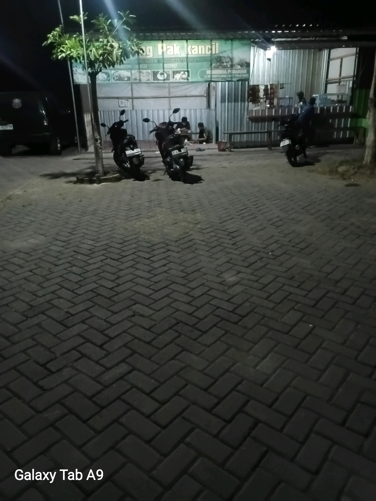 The image depicts an outdoor scene at night, likely in Bendorejo, Indonesia. In the foreground, a paved area with interlocking dark gray bricks is visible. The bricks are laid in a herringbone pattern, creating a textured surface.
In the mid-ground, several motorcycles are parked on the paved area. Some appear to be scooters, with their headlights and taillights off. In the background, a building with a metal facade and a green sign is present. The sign has text in Indonesian, and a portion of it reads "G Pak kancil". There are also some people in the background, some appearing to be seated at tables. A dark-colored vehicle, possibly a van, is parked on the left side of the image. A tree with green foliage stands near the left side of the building. The overall lighting suggests artificial illumination from the building and possibly streetlights. The scene appears to be a parking lot or an area in front of a commercial establishment at night.