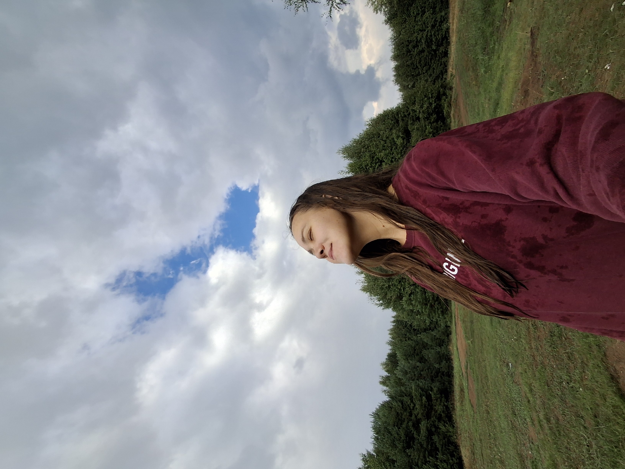 A young woman with long, dark hair, wearing a burgundy sweatshirt, stands in a field facing upwards with her eyes closed and a slight smile. The sweatshirt appears damp, with darker patches suggesting moisture. Behind her, a dense line of dark green coniferous trees forms a backdrop. The ground in front of the trees is a mix of green grass and patches of drier, reddish-brown earth. The sky above is filled with dramatic, gray clouds, but a small patch of bright blue sky is visible, with a thin wisp of cloud. The overall impression is one of enjoying a moment of fresh air outdoors. The location is described as Plishkino, Russia, suggesting a rural or semi-rural setting.