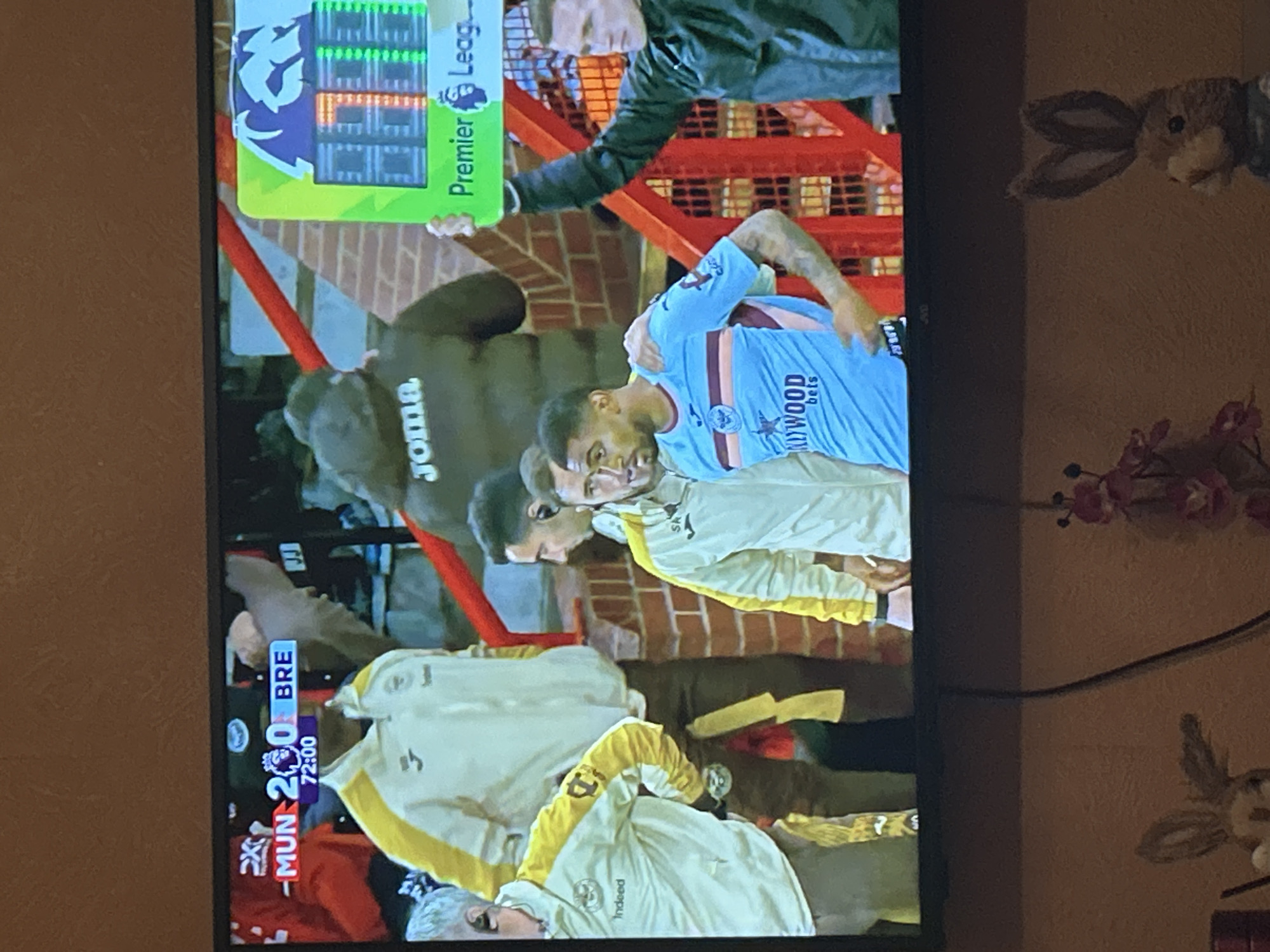 The image displays a television screen broadcasting a Premier League football match, observed in an indoor setting. The screen is filled with the ongoing game, showing several individuals on what appears to be the pitch or sideline area.
On the bottom left of the TV screen, a graphic overlay indicates the score: "MUN 2 - 0 BRE," with "72:00" showing the match time in the 72nd minute. The Premier League logo is visible on this scoreboard and also on a larger substitution board held up by a person in a dark jacket in the background.
Key figures on the screen include a football player with dark skin and short dark hair, wearing a light blue and dark blue striped jersey that prominently features "HOLLYWOOD bets" as a sponsor. Nearby, another individual is seen in a light yellow/cream long-sleeved top with yellow accents. A third person, possibly a coach or staff member with gray hair, is visible at the bottom of the screen, also in a similar light yellow/cream top with "Indeed" printed on the sleeve. In the mid-ground, a dark figure with "JOMA" written on their back is also visible. The background elements of the broadcast show typical stadium architecture, including red barriers and brick walls.
To the right of the television, on a plain wall with a warm, earthy tone, are decorative items. A plush or woven rabbit's head with large ears is mounted on the wall, and below it, a small cluster of artificial pink flowers adds a touch of color. A dark cable runs horizontally across the wall. The scene captures a casual moment of watching sports.