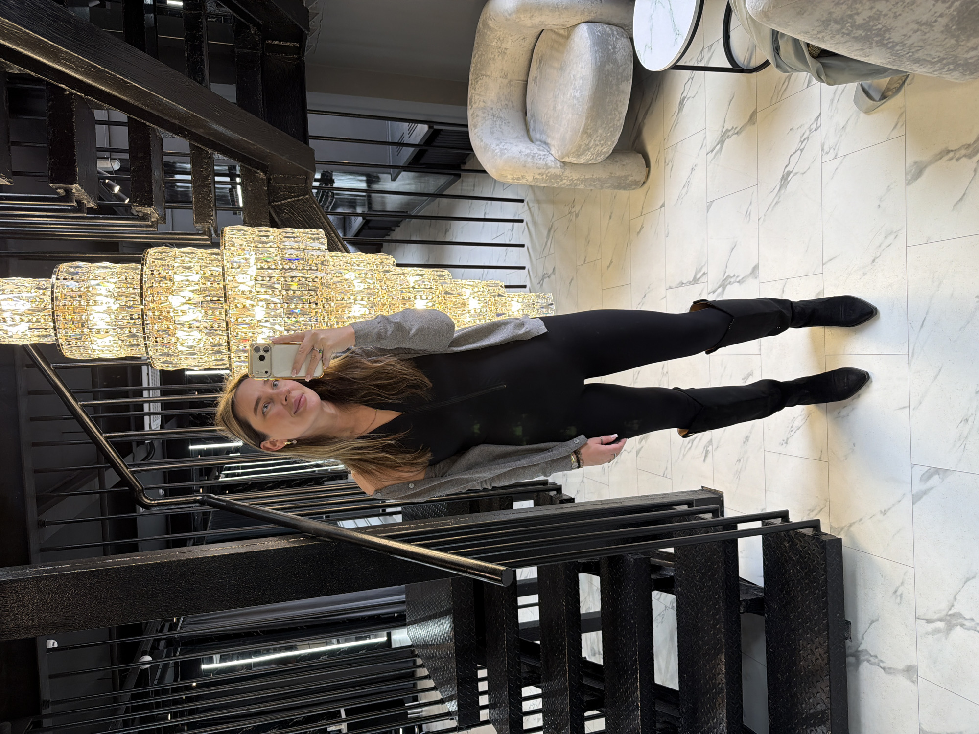 A woman stands inside a building in Moscow, Russia. She is taking a selfie with a smartphone in front of a modern staircase with a large crystal chandelier. She wears a gray sweater, black top, black leggings, and black heeled boots. The staircase is black metal. The floor is white marble with gray veins. There is a gray swivel chair visible in the background.