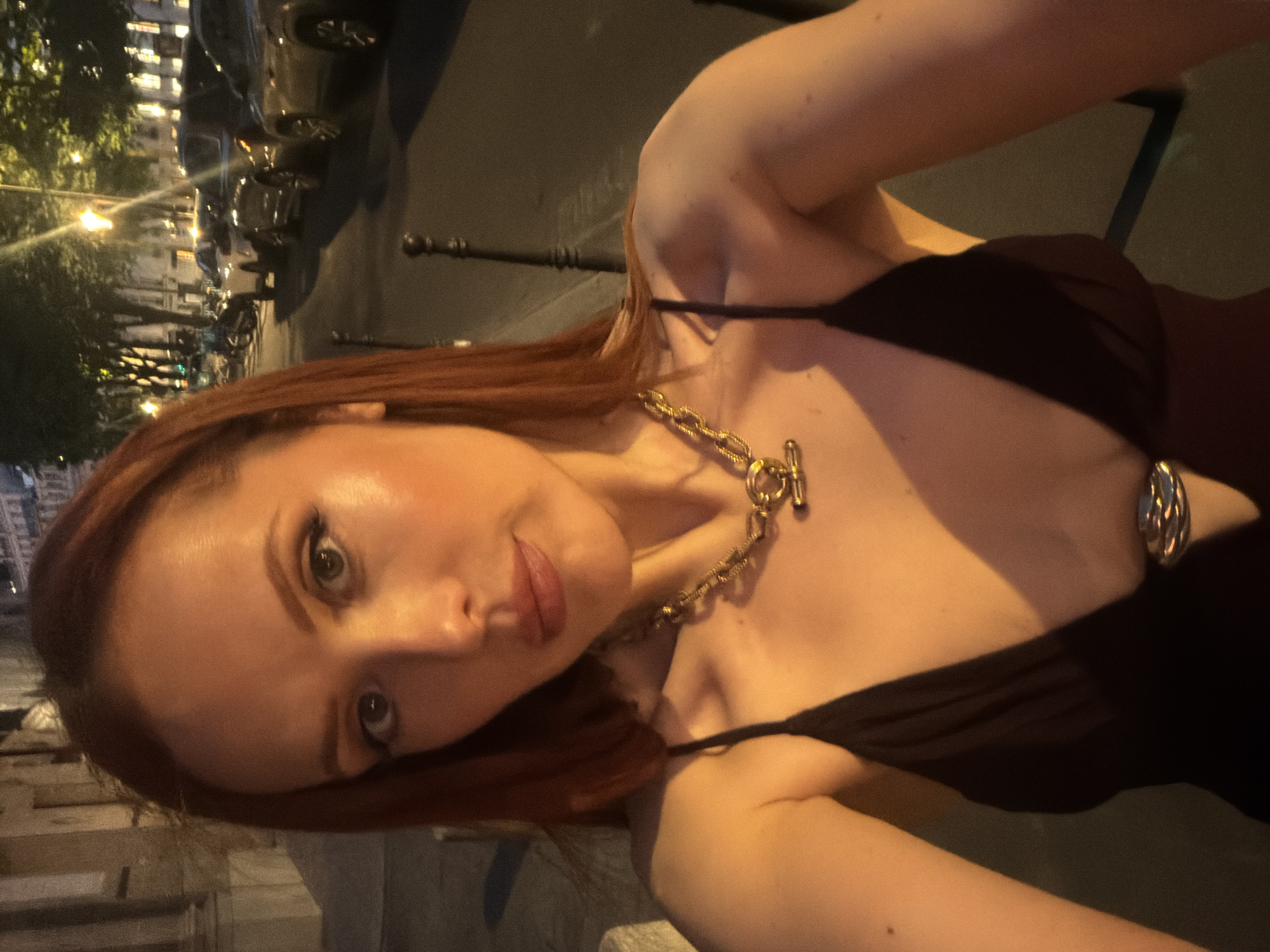 A woman with auburn hair and fair skin is posing for a selfie at night in Paris, France. She is wearing a dark-colored, low-cut dress with thin straps and a gold chain necklace. In the background, Parisian streets can be seen, lined with parked cars, buildings illuminated by streetlights, and wrought-iron railings. The scene is dimly lit, suggesting it is late at night.
