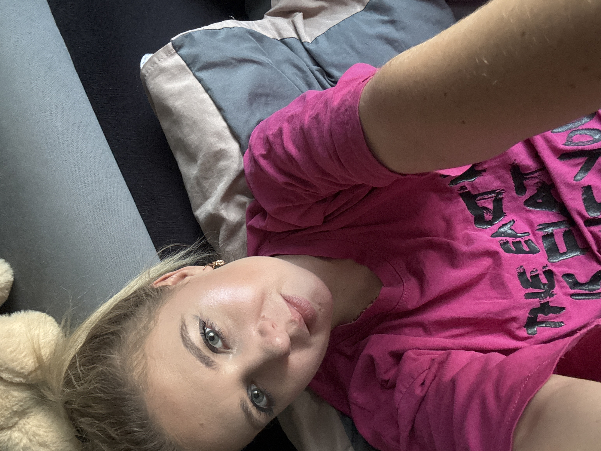 A close-up, overhead, selfie-style shot shows a young white woman with blonde hair pulled into a high bun. She is lying down and wearing a bright pink t-shirt with black lettering that appears to say "THE EAST IS..." and then some illegible text below. She has on light makeup, including winged eyeliner and lip gloss, and is wearing a small gold earring. Her left arm is visible, bent at the elbow. She is positioned on a bed with a striped pillow that has tan and grey sections. The background appears to be a grey upholstered headboard. The lighting is bright, and the woman's skin has a dewy appearance. The scene is indoors, and the location within the city is unknown.