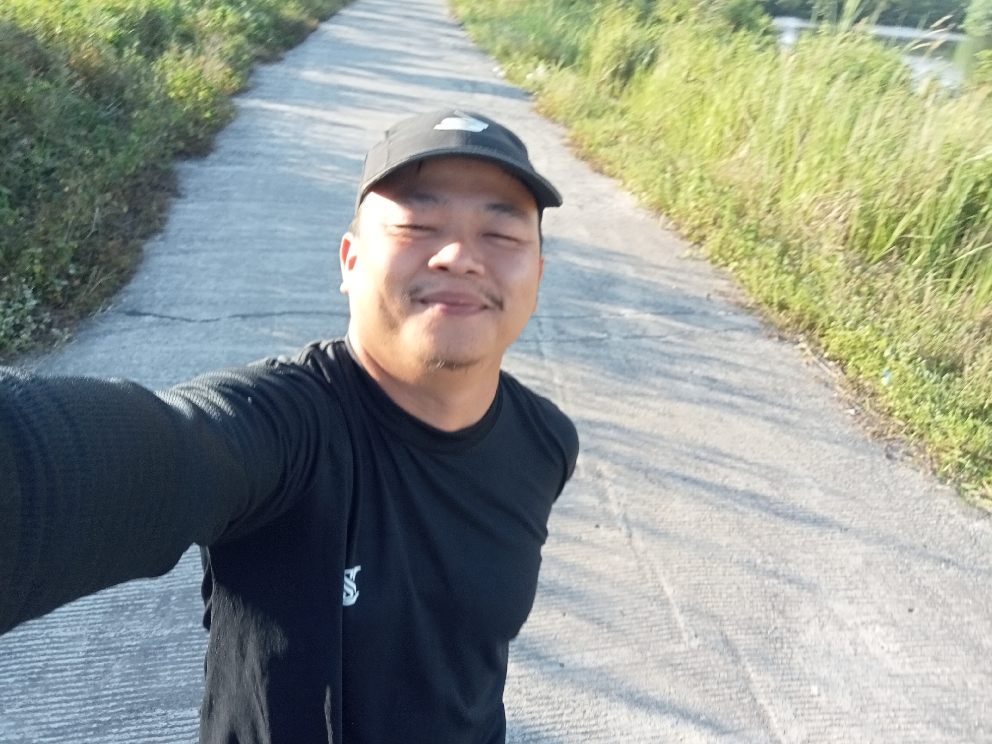 A male individual is taking a selfie outdoors on a paved road lined with tall grass. He is wearing a black baseball cap and a black t-shirt. His eyes are closed, and he has a slight smile on his face. The road appears to be a rural or suburban area, potentially in the city of Kualu, Indonesia, given the lush vegetation and the style of the road. The lighting suggests it is daytime, possibly late afternoon due to the long shadows cast by the grass. The man's left arm is extended, holding the camera or phone. The background shows a receding road and a dense growth of green grass on either side.