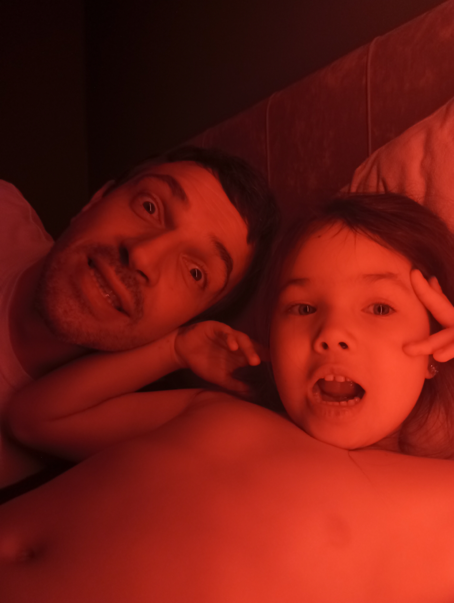 A photograph depicts an adult male and a young girl lying down, possibly in bed, under red lighting. The man is on the left side of the frame, with his face close to the camera. The girl is on the right, with her mouth open. The photograph is taken from a close-up angle. The setting lacks specific contextual clues to indicate a location in Pervomaysky, Russia.