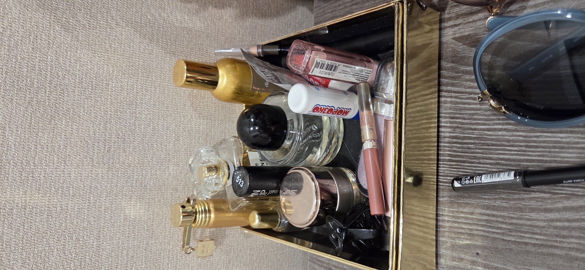 The image shows a golden tray filled with various cosmetics and perfumes on a wooden surface. The items include several perfume bottles, one with a clear faceted cap and another with a gold cap, a black lipstick tube, a pink lipstick, a lip balm with Russian labeling, a small glass bottle with a gold cap, and a transparent bottle with a black cap. To the left of the tray, a black hair clip is visible. A black eyeliner pencil and a pair of blue sunglasses are placed on the wooden surface in front of the tray. The background is a textured beige wall. The scene does not provide any specific information about the city of Lipetsk, Russia.
