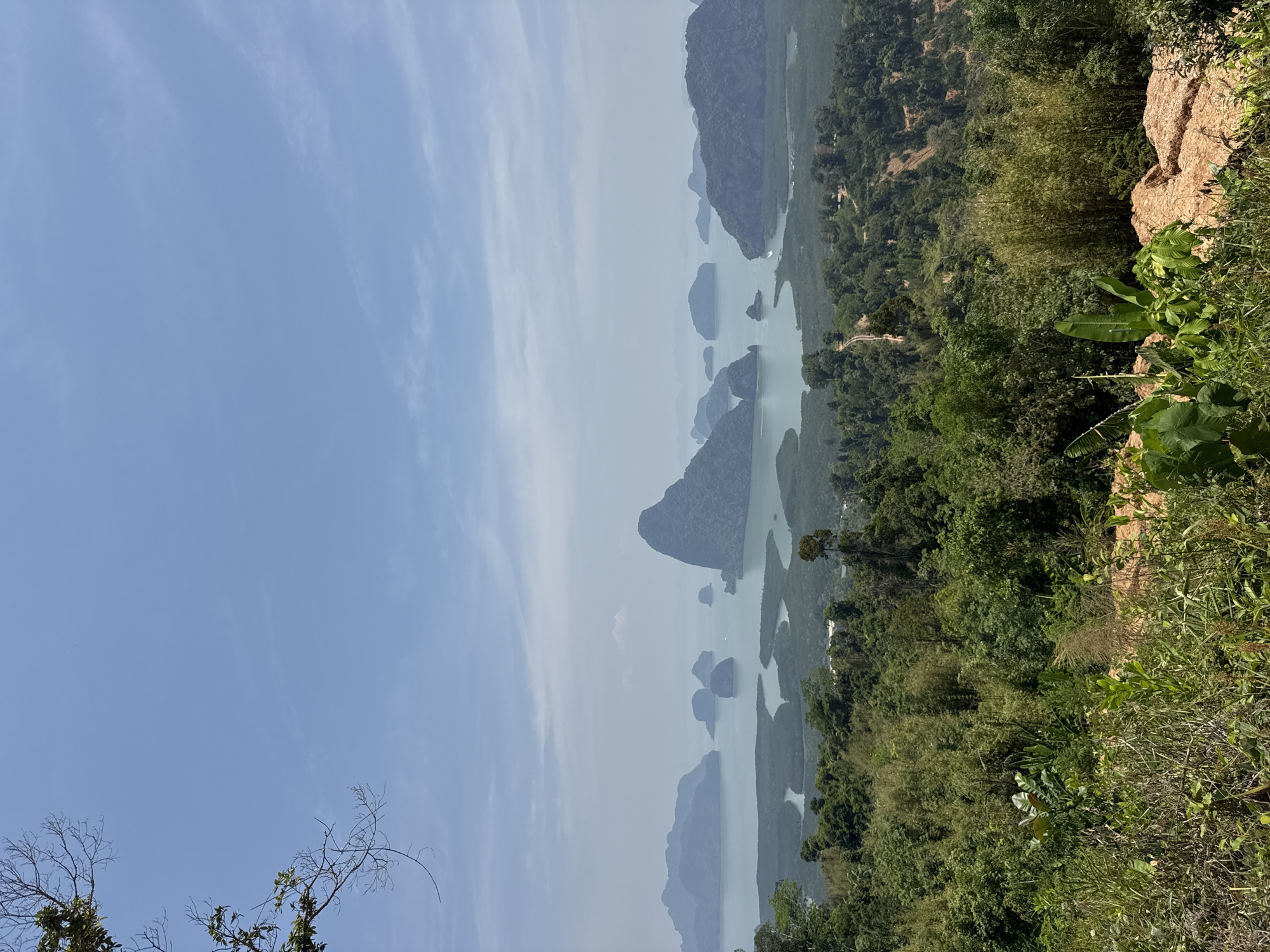 The image shows a landscape from an elevated perspective in Ban Khlong Khian, Thailand. The view features islands in a bay, interspersed with waterways. Lush vegetation covers the islands and extends into the foreground. The sky is light blue with wispy clouds. A rocky outcrop is visible on the right edge of the image.