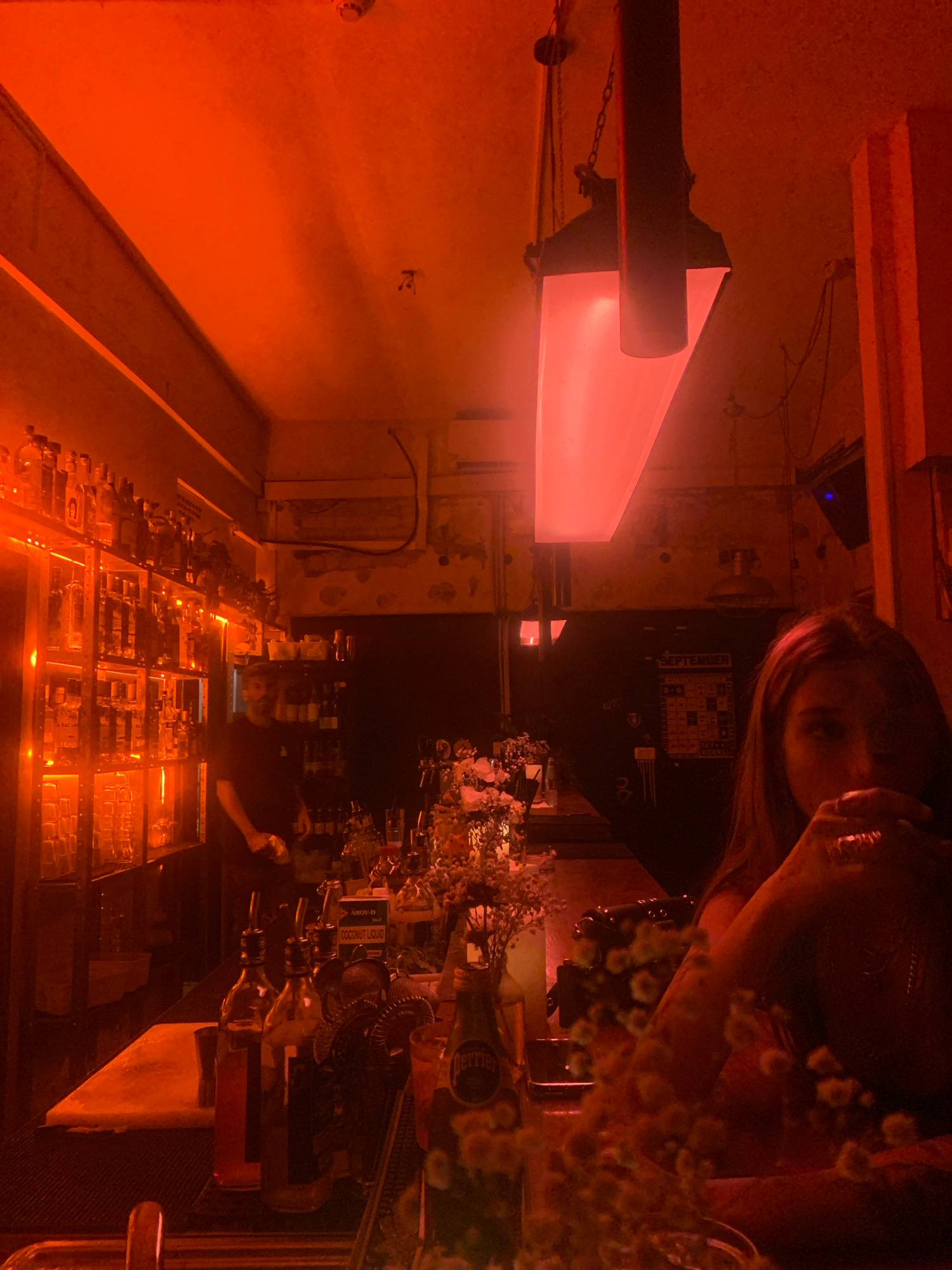 The image depicts the interior of a bar, dimly lit by red ambient lighting, with a focus on the bar counter and shelves stocked with bottles. On the left, a well-lit shelf unit displays numerous bottles of alcohol, with a person in a dark t-shirt visible behind the counter, appearing to be a bartender. The counter itself is made of a light-colored material, and several bottles, possibly containing syrups or mixers, are present. In the foreground, a woman is partially visible on the right, holding a drink and wearing rings on her fingers. The background includes a calendar labeled "SEPTEMBER" and a large red light fixture hanging from the ceiling. The overall atmosphere suggests a lively, perhaps late-night bar setting. This image is located in Tel-Aviv, Israel.