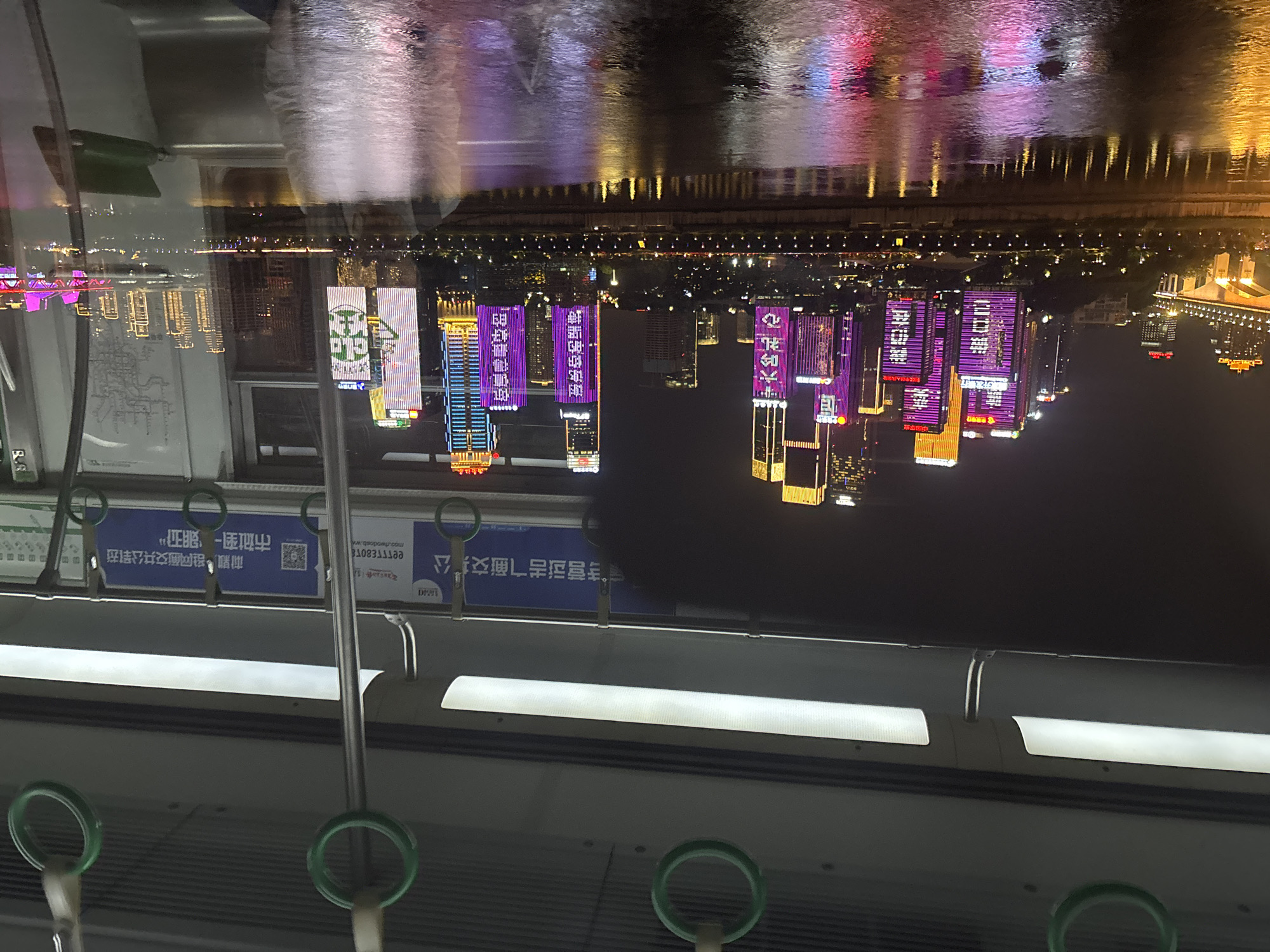 The image, taken from the inside of a train or subway car, shows a night view of what appears to be a skyline reflected on water. The cityscape, likely in Yuzhong District, China, based on the Chinese characters on nearby signs, displays numerous buildings with illuminated advertisements and designs, some colored purple, gold, and blue.
The interior of the train features overhead lighting, handholds, and informational signage. The focus is through the train window, capturing the urban scene and its reflections on a body of water, potentially a river or bay. The reflections create a distorted and inverted image of the city's lights.