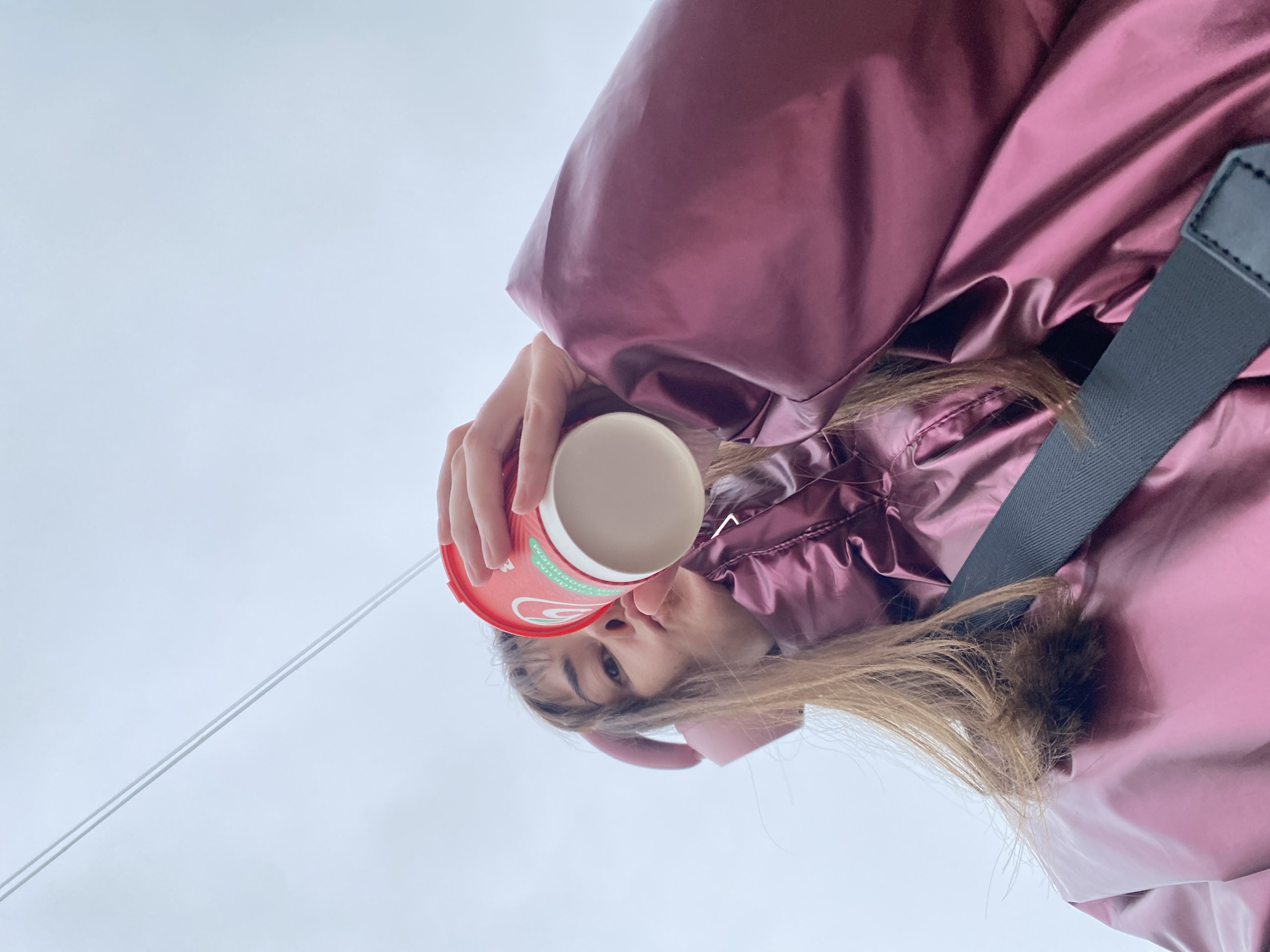 A person holds a red and white coffee cup towards the viewer, positioned at an upward angle. The individual wears a shiny, maroon-colored puffer jacket and has brown hair. A black strap crosses the body, visible near the jacket. The background is a muted, light gray, suggesting an overcast or cloudy day. A thin, white line extends into the sky. The location is indeterminable without additional cues, but the request is the city of Votkinsk, Russia.