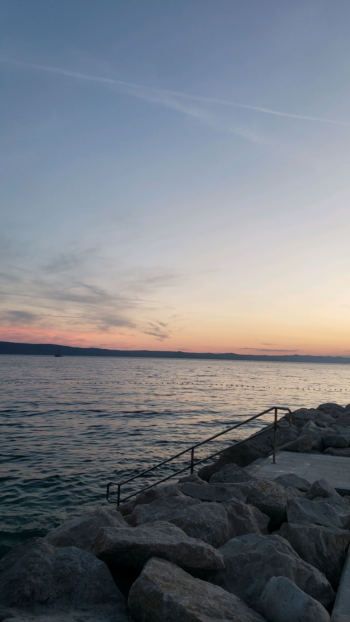 This image depicts a serene coastal scene at dusk in Općina Tučepi, Croatia. The horizon is marked by a distant, mountainous coastline silhouetted against a sky transitioning from a soft blue to hues of pink and orange. Wispy clouds are scattered across the upper atmosphere. In the middle distance, a lone sailboat is visible on the calm, dark water. A line of buoys or markers stretches across the sea. The foreground is dominated by a rocky shoreline, with large, gray stones forming a natural breakwater. A metal railing with accompanying steps leads down towards the water from a concrete pathway at the top of the rocks. The water exhibits gentle ripples. The overall atmosphere is tranquil and natural.