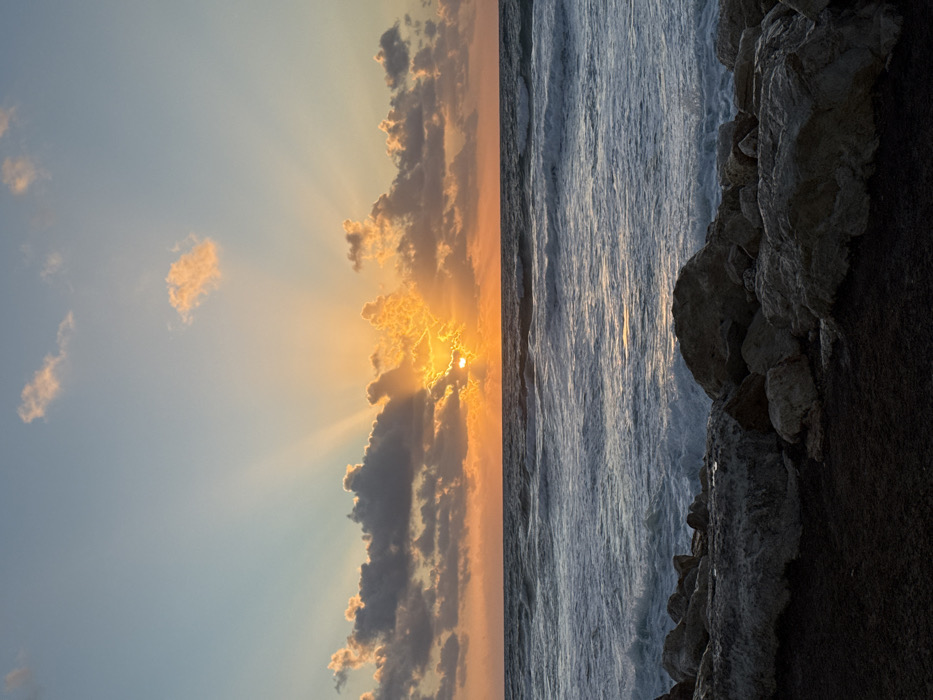 The image displays a coastal scene at sunset, likely along the Tel Aviv coastline in Israel. The sun is positioned low on the horizon, partially obscured by a band of dark, horizontally elongated clouds. Intense golden-orange light radiates from behind these clouds, forming distinct crepuscular rays that ascend into the upper sky. The upper sky transitions from a soft orange-yellow near the horizon to a pale blue at the top of the frame, with scattered, lighter clouds.
The midground is dominated by the Mediterranean Sea, characterized by moderate waves with white caps and foam, reflecting a subdued golden sheen from the setting sun on their crests. The water appears dark blue-grey overall.
In the foreground, a rocky shoreline is visible. Large, irregular grey and brown rocks are piled along the water's edge, some appearing wet from the waves. Below the rocks, a strip of dark, possibly damp earth or coarse sand, occupies the bottom of the frame. No individuals, animals, or man-made structures are discernible in the scene.