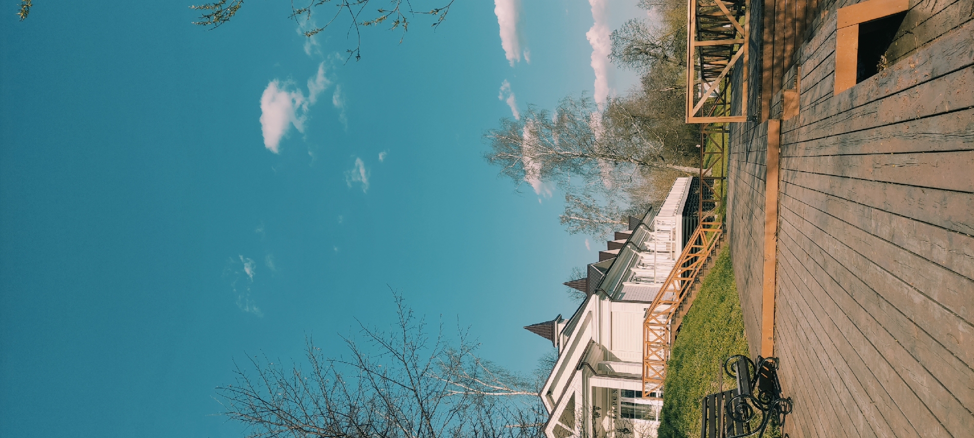 The image captures a scene in Grebnevo, Russia. A white, two-story building with a brown roof and decorative gables stands on a grassy lawn. The building is partially obscured by leafless trees in the foreground. A wooden boardwalk with a brown railing extends to the right of the building, leading out of frame. The sky is a clear blue with scattered white clouds. A portion of a bicycle is visible near the building.