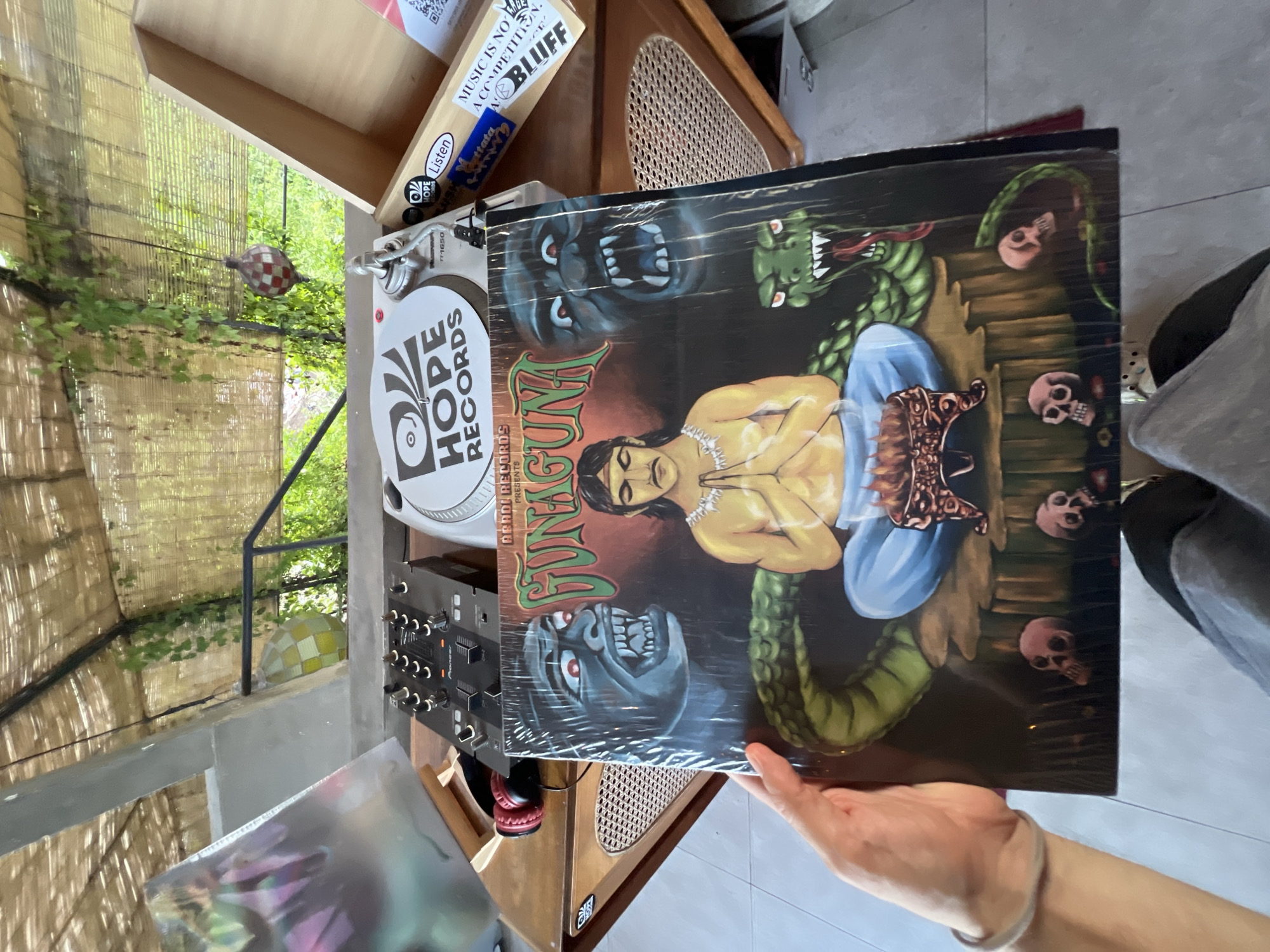 A close-up, slightly tilted image captures a person's hands holding a vinyl record, presented in what appears to be an outdoor or semi-outdoor music-themed setting in North Kuta, Indonesia. The album cover features a striking illustration: a central figure with dark hair and bare chest, sitting in a meditative pose with clasped hands. Around this figure, a large green snake with red eyes and fangs coils, and several pink skull-like objects are scattered on a wooden platform. On the left side of the cover, a blue-grey monster with glowing red eyes and sharp teeth looms. The album's title, "FUNGUN," is prominently displayed in green, stylized lettering, with "ABADI RECORDS PRESENTS" visible below it. The record is encased in clear plastic shrink wrap.
To the left of the record, a white turntable is partially visible, featuring a circular sticker with the "HOPE RECORDS" logo. Below the turntable, a black Pioneer audio mixer with numerous knobs and faders is integrated into a wooden console. The background shows lush green foliage and bamboo or woven screens, suggesting a tropical, relaxed environment, illuminated by natural daylight. A small, checkered, gourd-like decorative object hangs in the mid-ground.
On a wooden shelf behind the turntable, several visible stickers and labels include text such as "MUSIC IS NO A COMPETITION. AYE BLUEF," "Listen," "Guttata," "1650," and another "HOPE" logo. The image conveys a casual, music-centric atmosphere, likely a record store or cafe, where vinyl records are displayed and enjoyed.